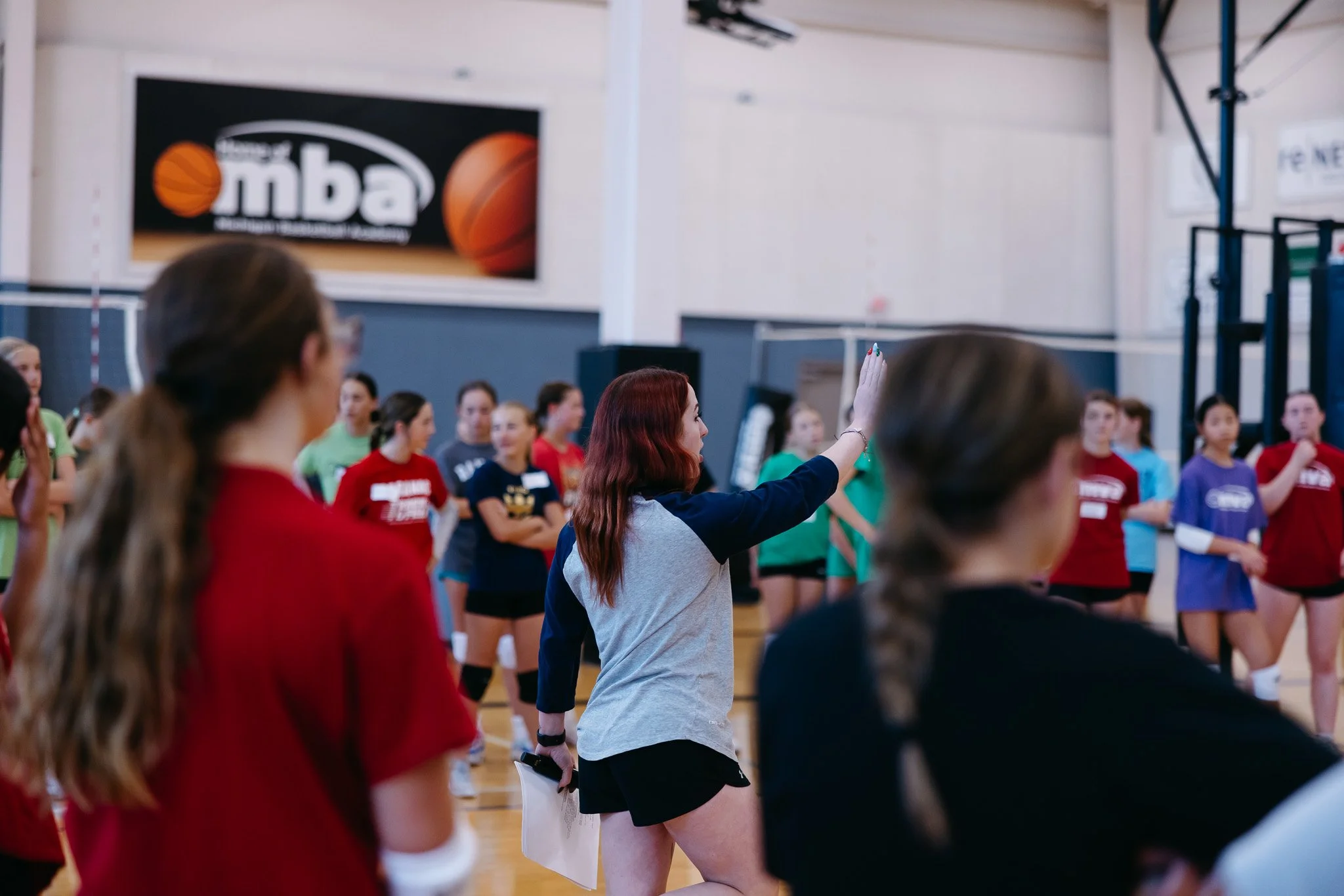 A group of young women standing in a gymnasium, participating in a basketball event. One woman with red hair, in a gray sweatshirt and black shorts, is raising her hand, possibly asking or answering a question.