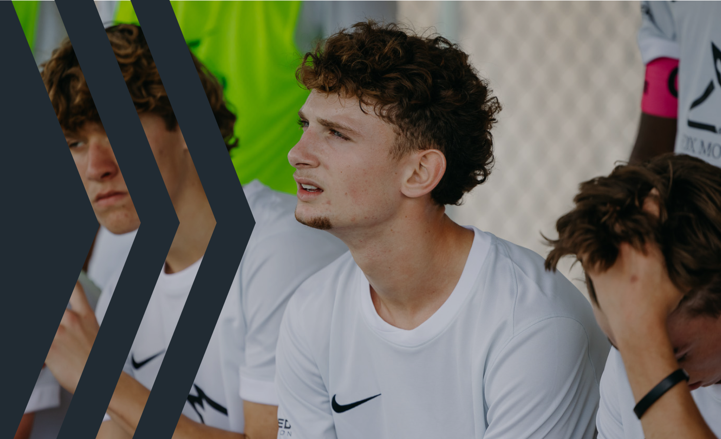 Close-up of a young man with curly brown hair and a light beard, wearing a white Nike sports shirt, sitting with others in a sports setting, showing a concerned or focused expression.