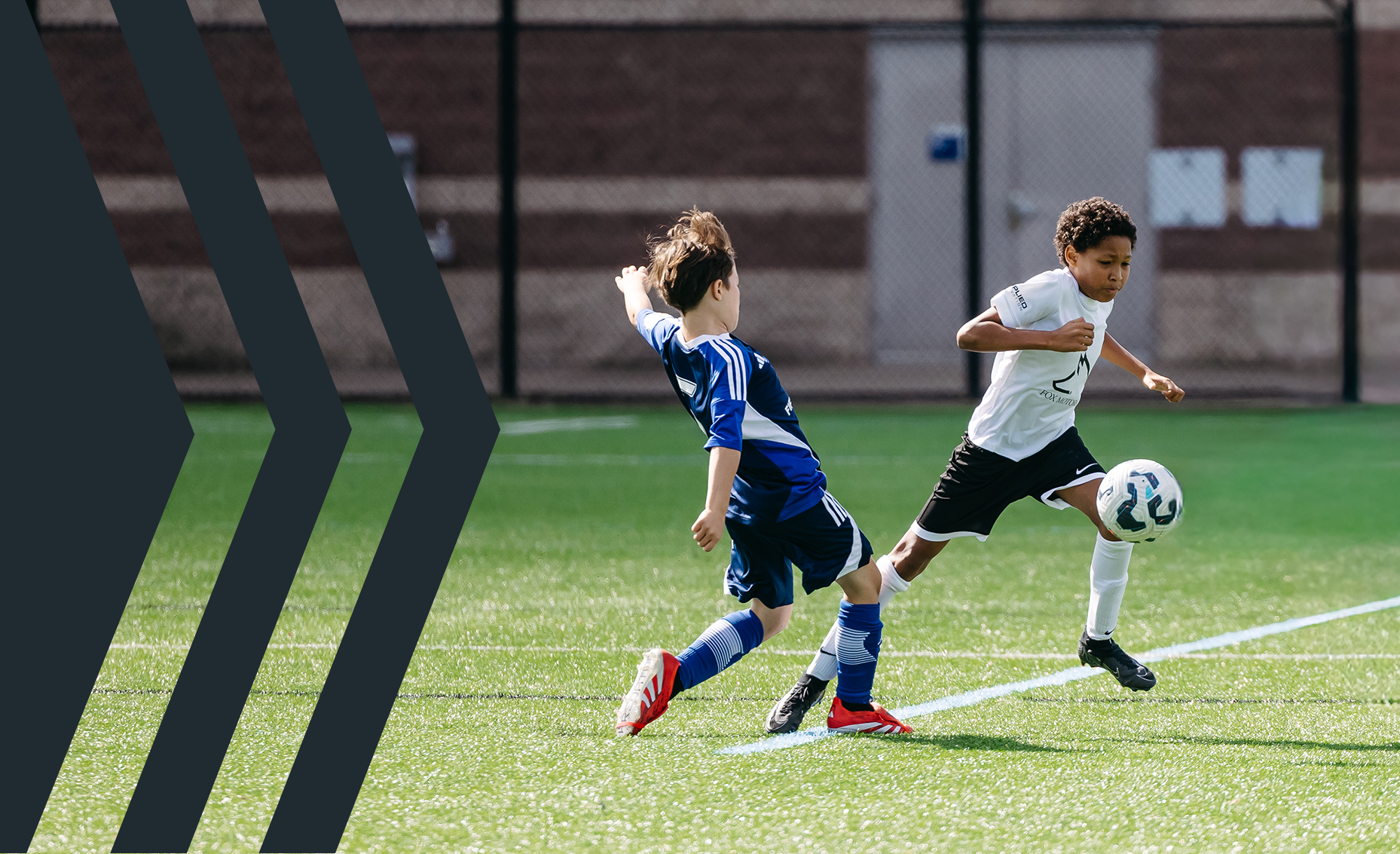 Two young boys playing soccer on a field, one in a white shirt and black shorts kicking the ball, the other in a blue jersey and shorts approaching the ball.