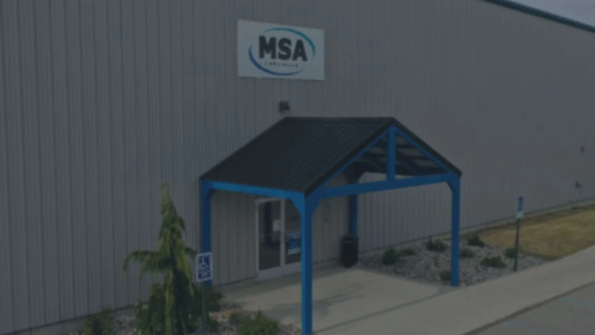 Exterior of a large industrial or commercial building with a sign reading 'MSA' on the wall. There is a small blue entryway with a roof and a glass door, surrounded by gravel and some small plants.