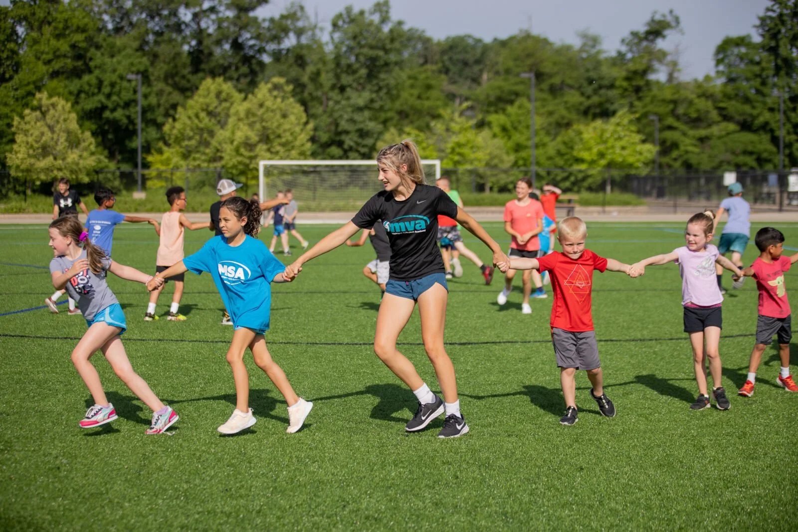 Children holding hands and playing in a circle on a grassy field, instructor or coach among them, in a park or sports field with trees and a goal in the background.