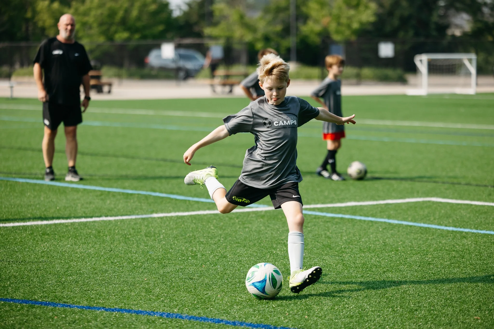 A young boy playing soccer on a green field during daytime, with a coach and other children in the background.
