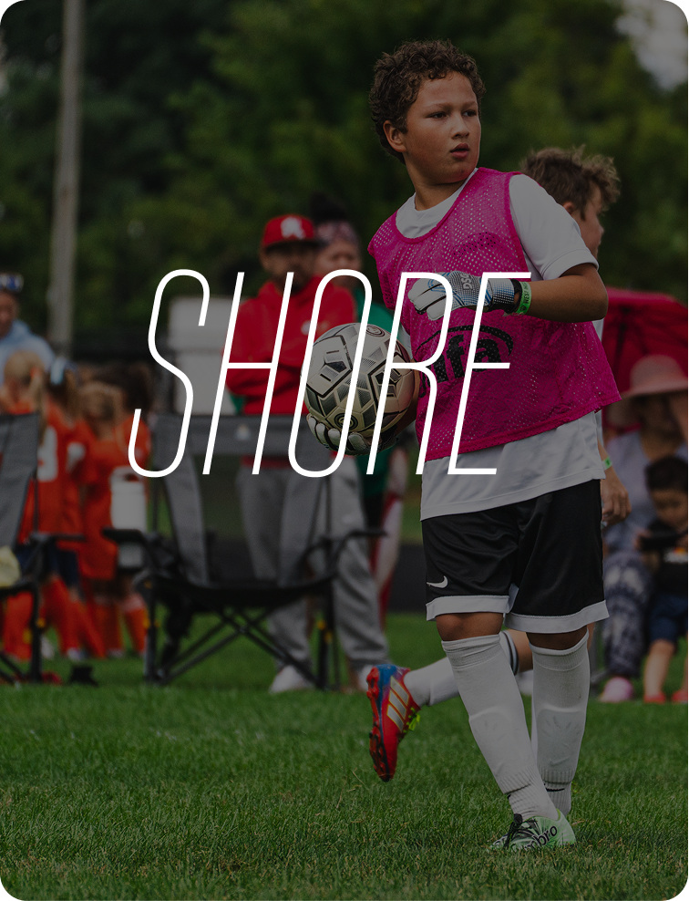 Young boy in a soccer uniform holding a soccer ball during a game or practice, with a crowd of people watching in the background, outdoors on a grassy field.