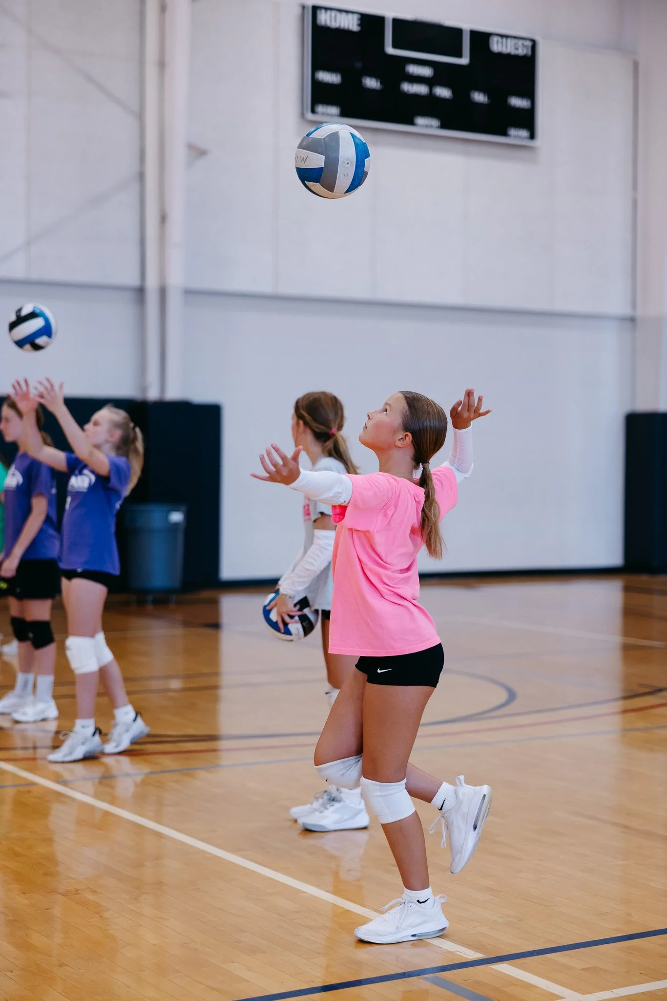 Young girls participating in a volleyball practice or game in an indoor gym, with volleyballs, wearing athletic clothing, including knee pads and sneakers.