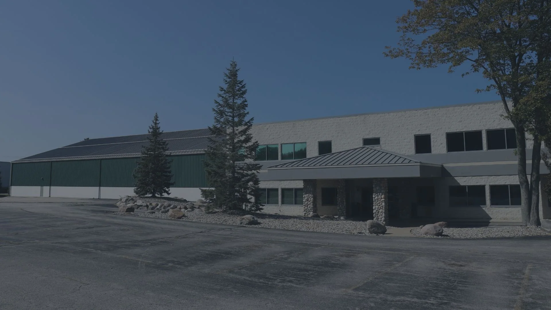 A modern commercial building with a mix of stone and metal siding, surrounded by trees and rocks, and an empty parking lot in front.