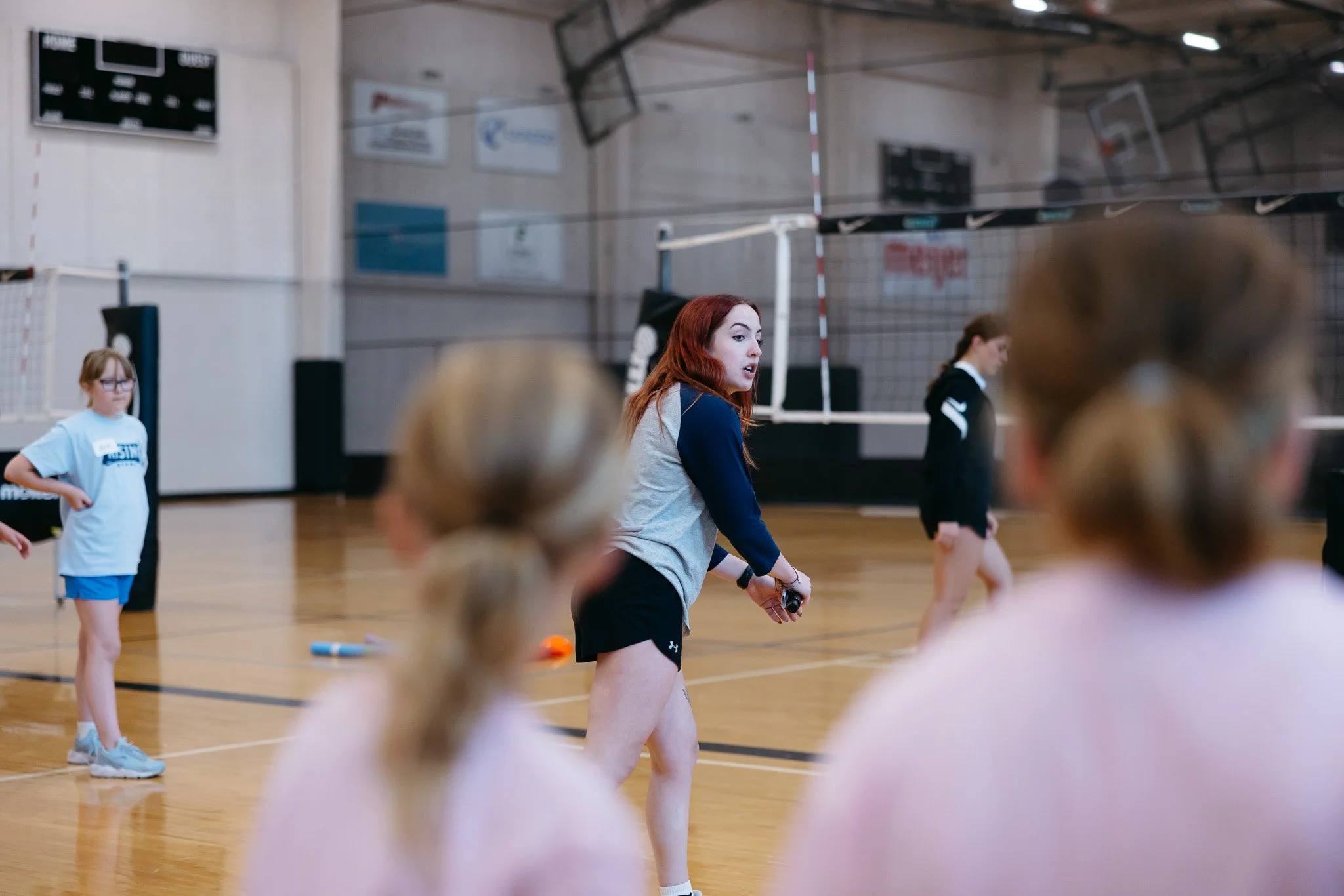 A group of children and a young woman in a gymnasium, possibly participating in a sports or physical activity session. The young woman appears to be instructing or coaching, and some children are wearing sports uniforms.