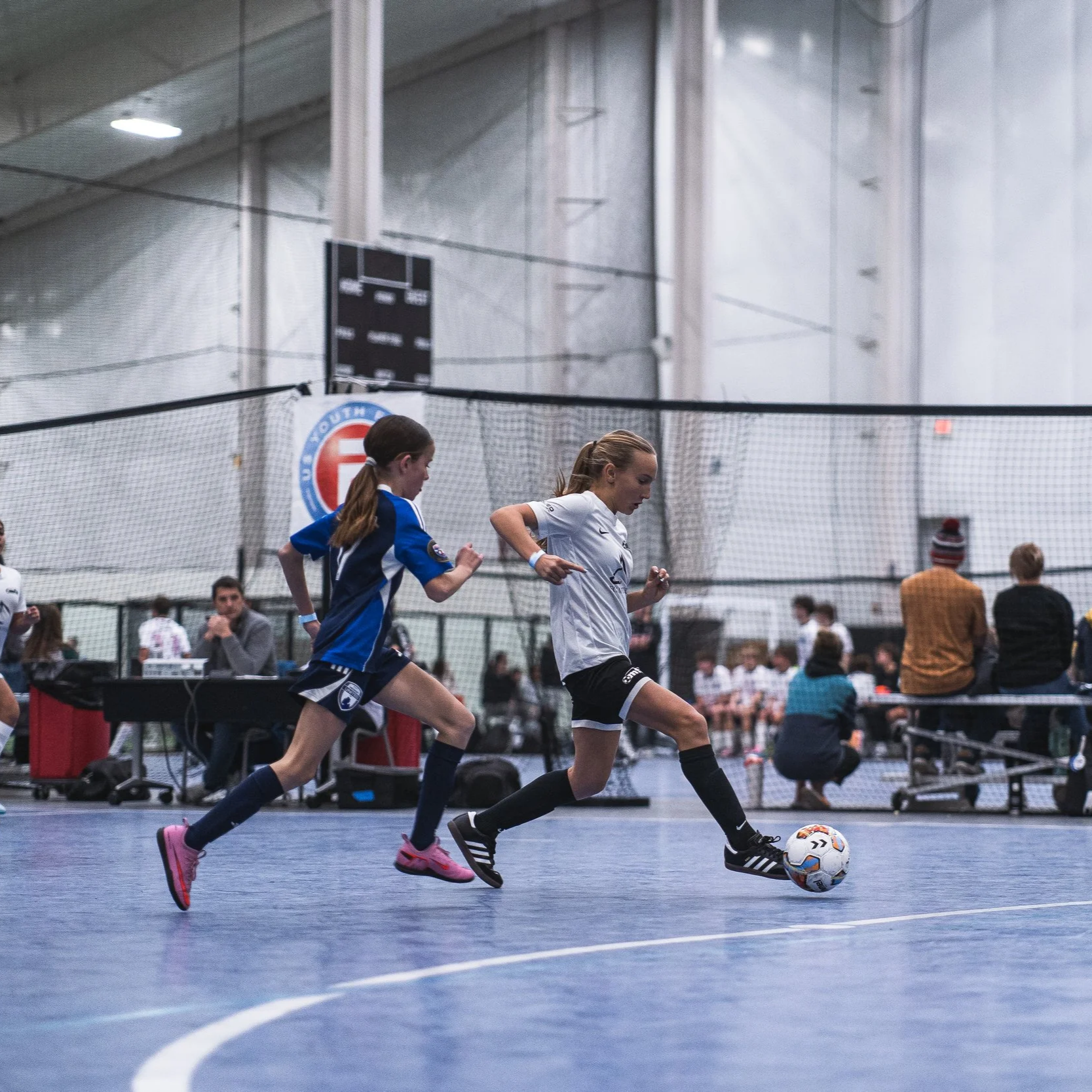 Two young female soccer players in indoor sports facility competing for control of a soccer ball. One player wearing a white jersey and black shorts, the other wearing a blue jersey and blue shorts. Spectators and other players in the background.