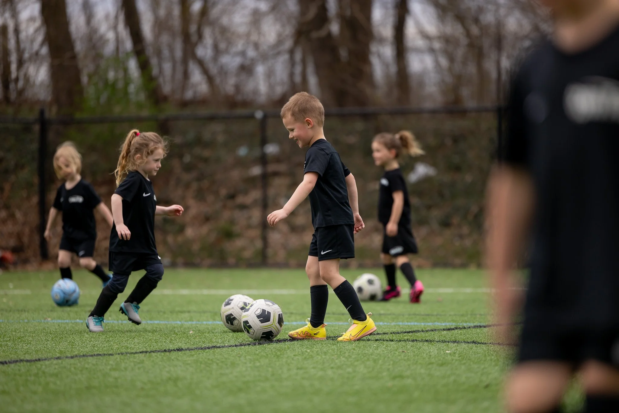 Young children playing soccer on a field, kicking soccer balls, with trees in the background.