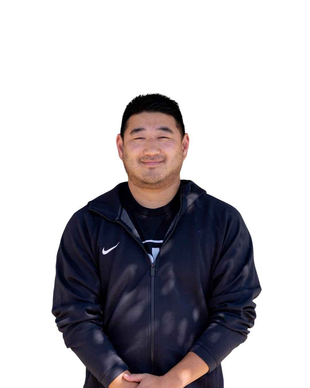 A smiling man with short black hair and facial hair, wearing a black Nike jacket and black T-shirt, standing with arms crossed, against a white background.
