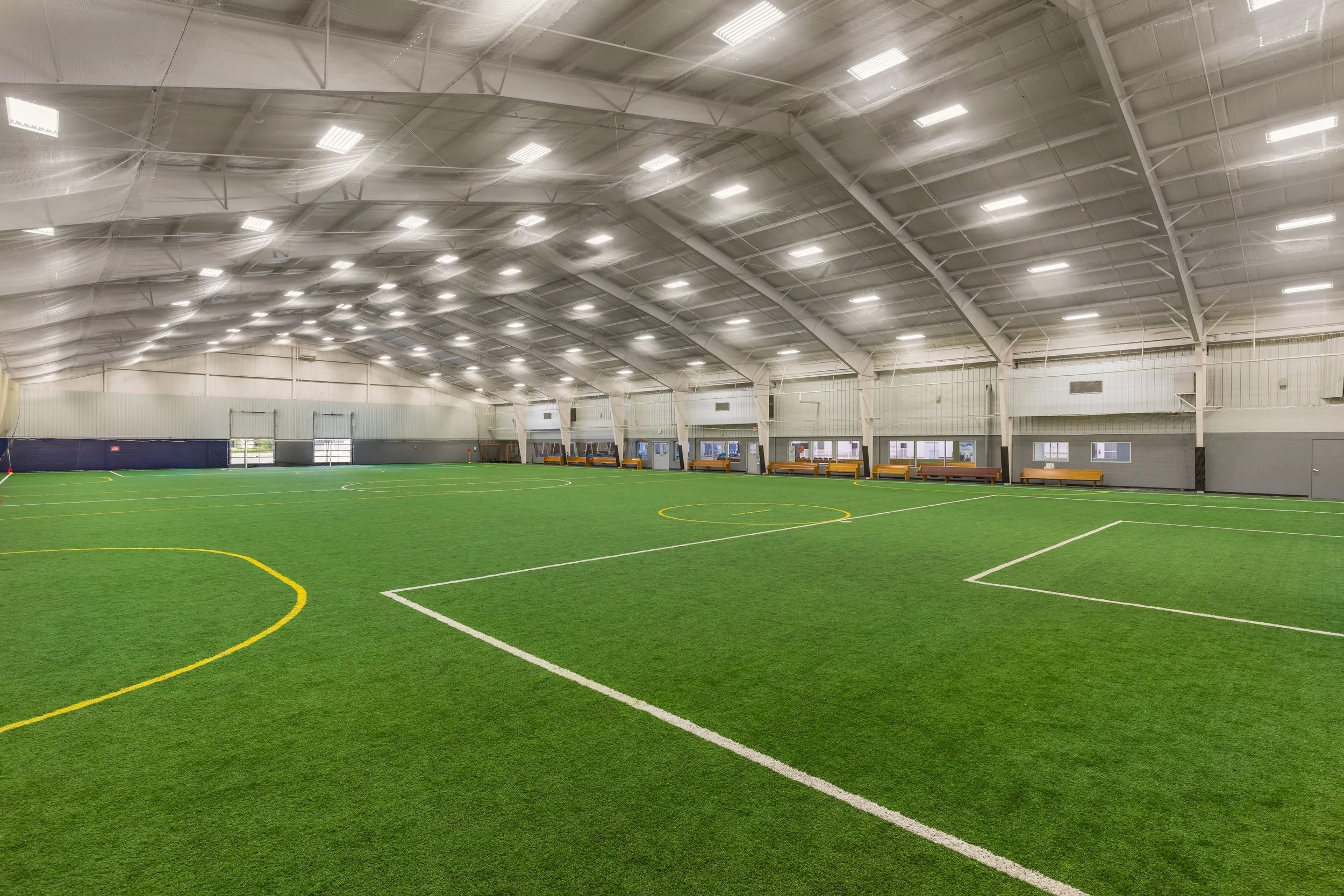 Indoor sports facility with a large, green artificial turf field and a high, curved ceiling with bright overhead lights.