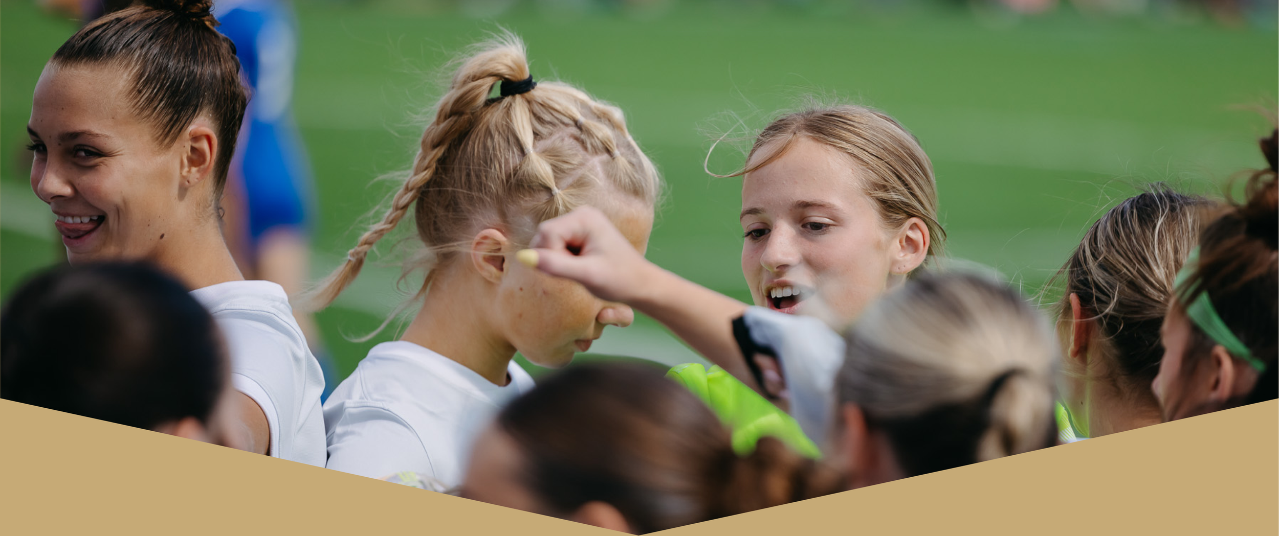 Young women and girls in sports uniforms gather on a field, some with braided hair, as a woman applies face paint or makeup on one of the girls' faces.