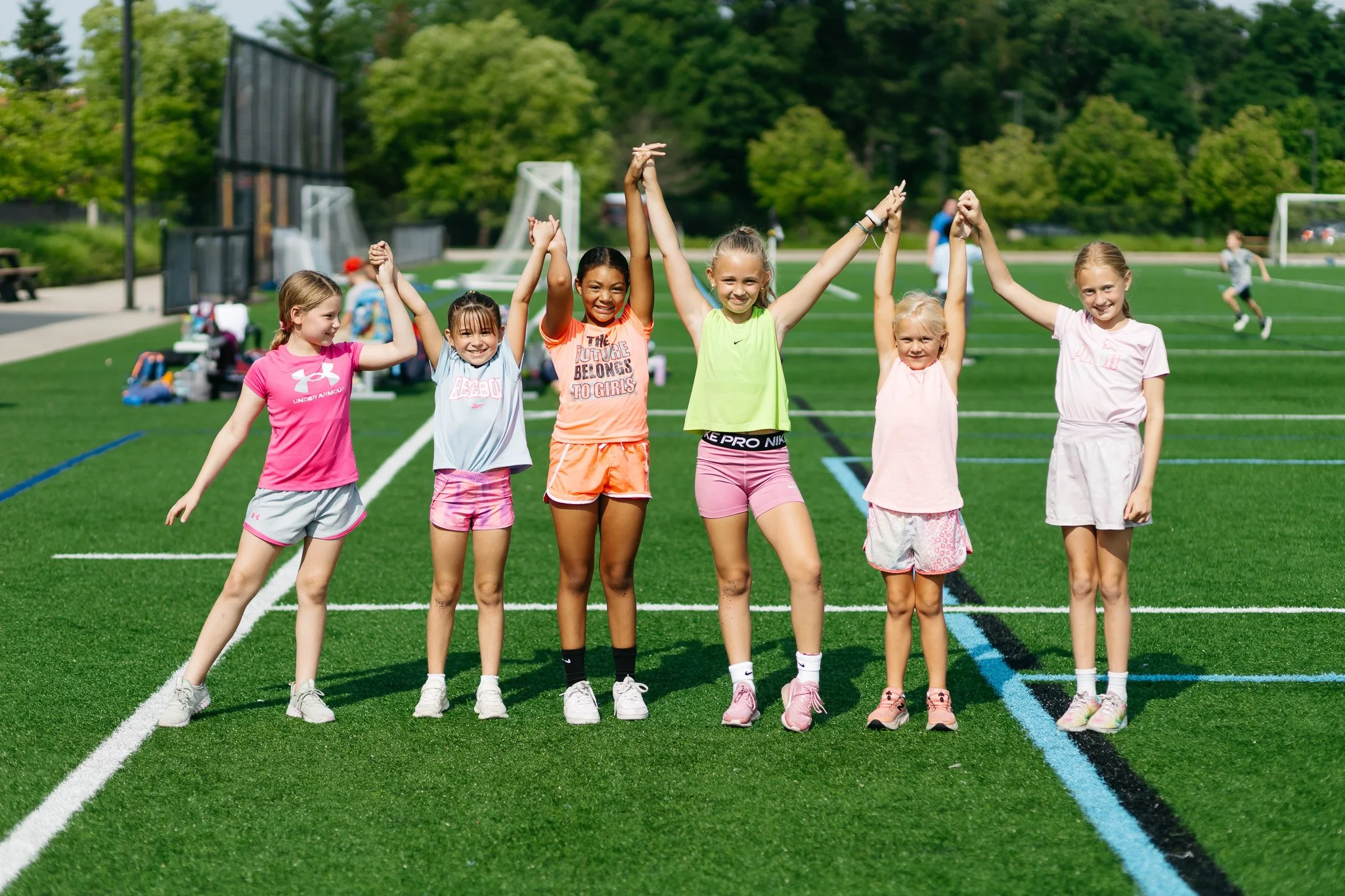 Group of five young girls standing in a row on an outdoor sports field, holding hands and smiling, surrounded by other children playing in the background on a sunny day.