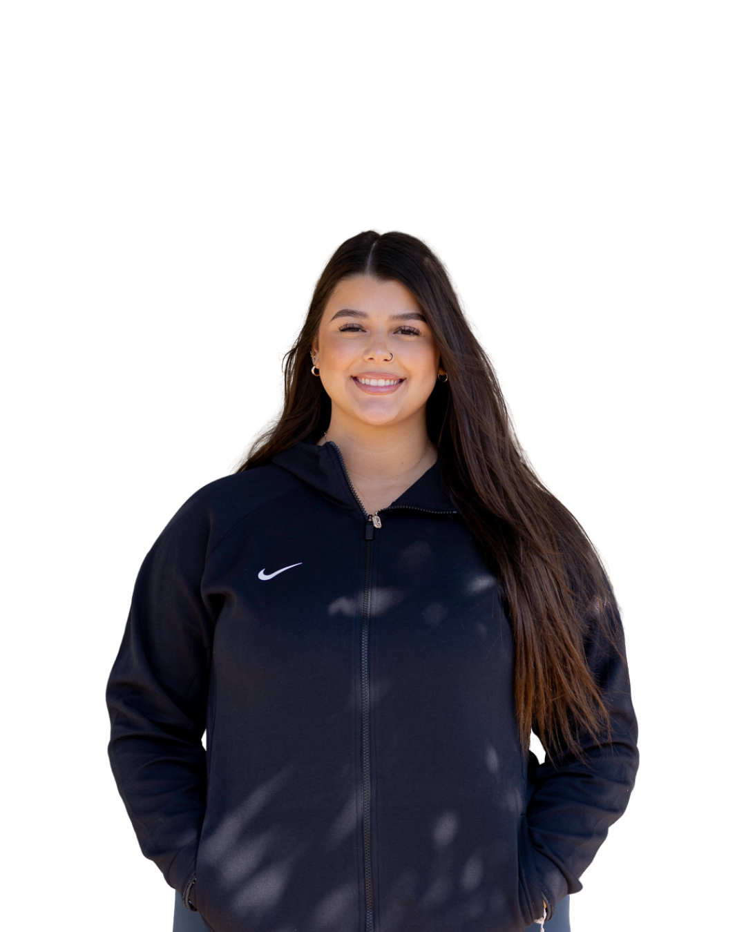 A young woman with long brown hair smiling, wearing a black Nike zip-up jacket with her hands in her pockets, standing in front of a white background.