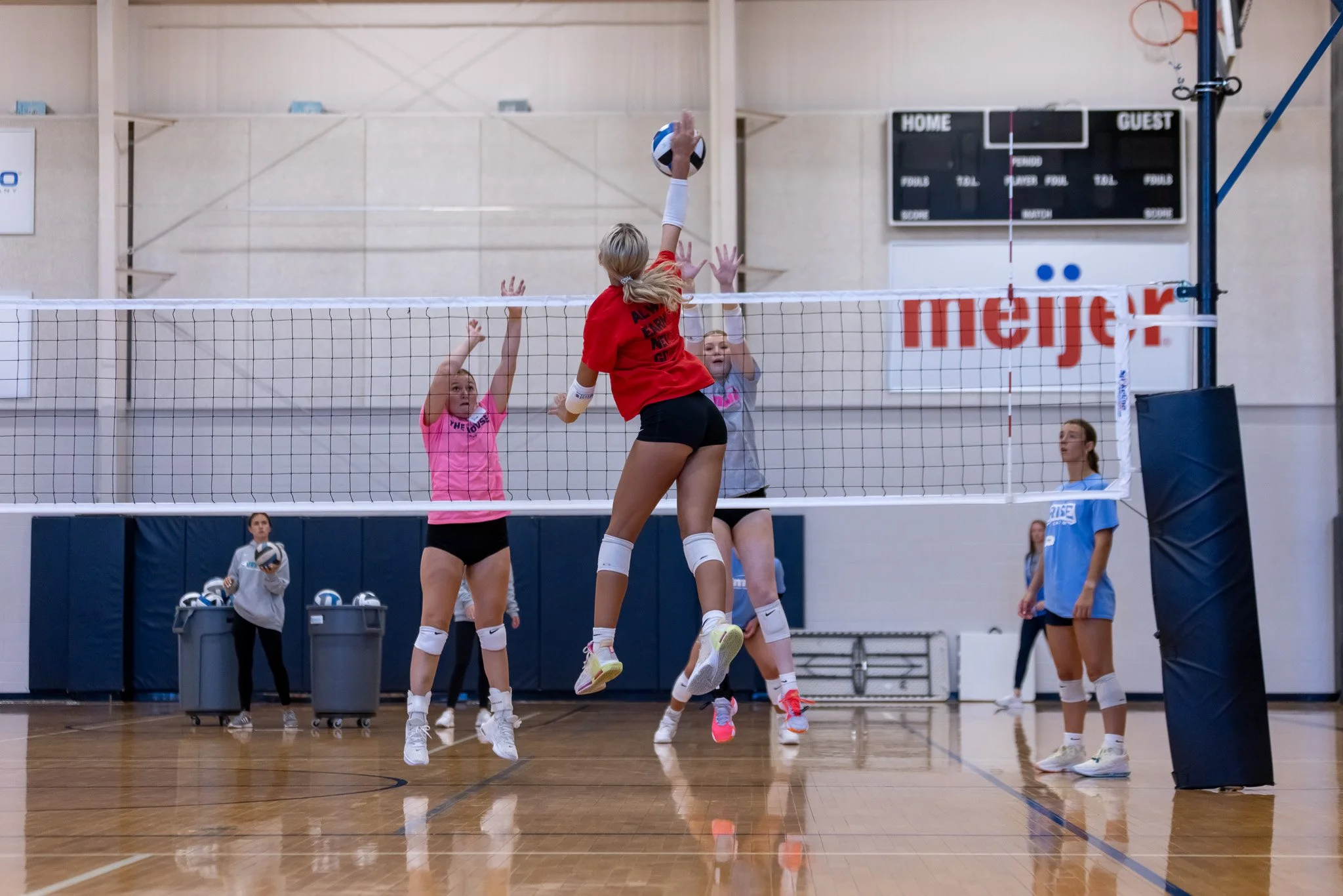 A volleyball game is taking place inside a gymnasium, with a female player in a red shirt jumping to hit the ball at the net. Two other female players in pink and light blue shirts are attempting to block or receive the ball. Several other players and a referee are visible in the background.