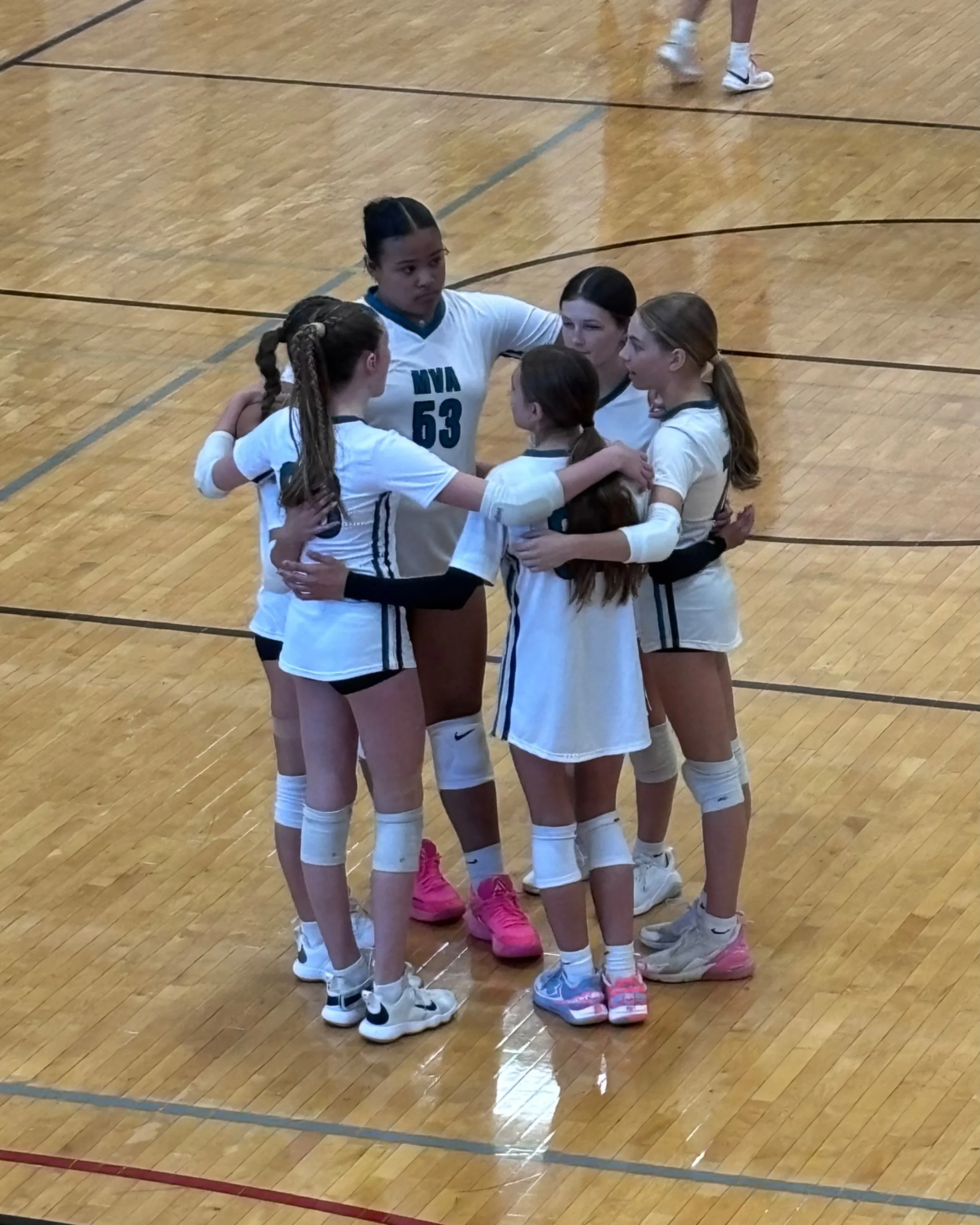 A group of five young women in volleyball uniforms standing in a circle indoors, holding each other in a team huddle on a wooden gymnasium floor.