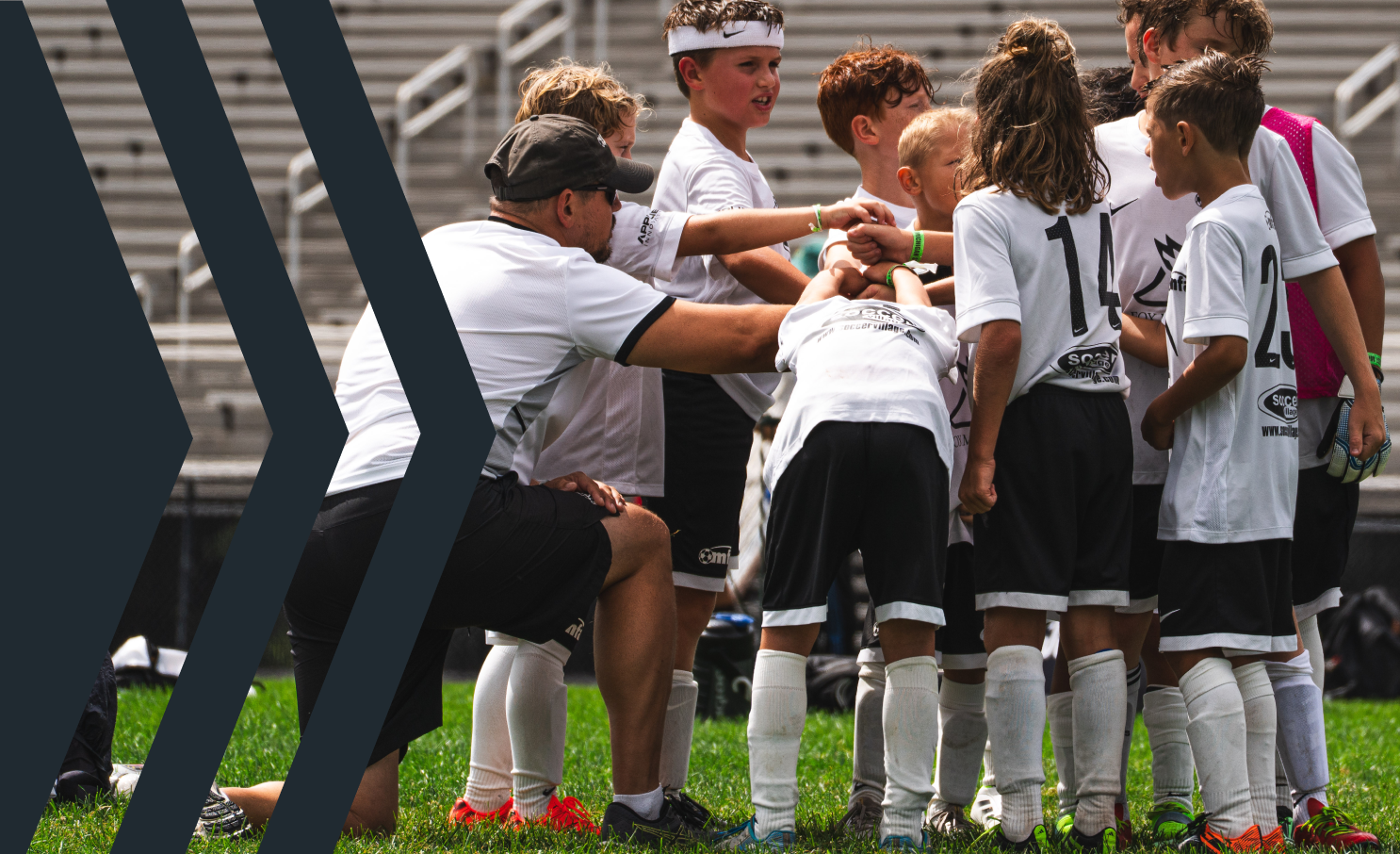 Youth soccer team in a huddle with coach during a game, players wearing white jerseys and black shorts on the field.