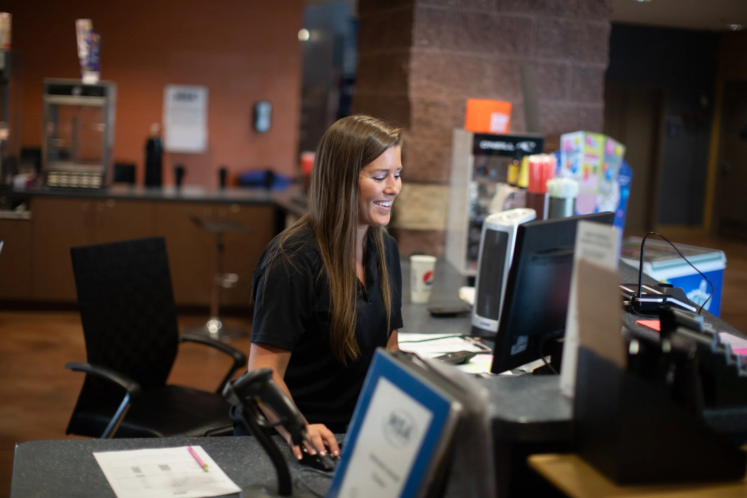 A woman with long brown hair sitting at a desk in an office or reception area, smiling while working on a computer with multiple monitors. The background features a brick wall, colorful products, and a coffee cup.