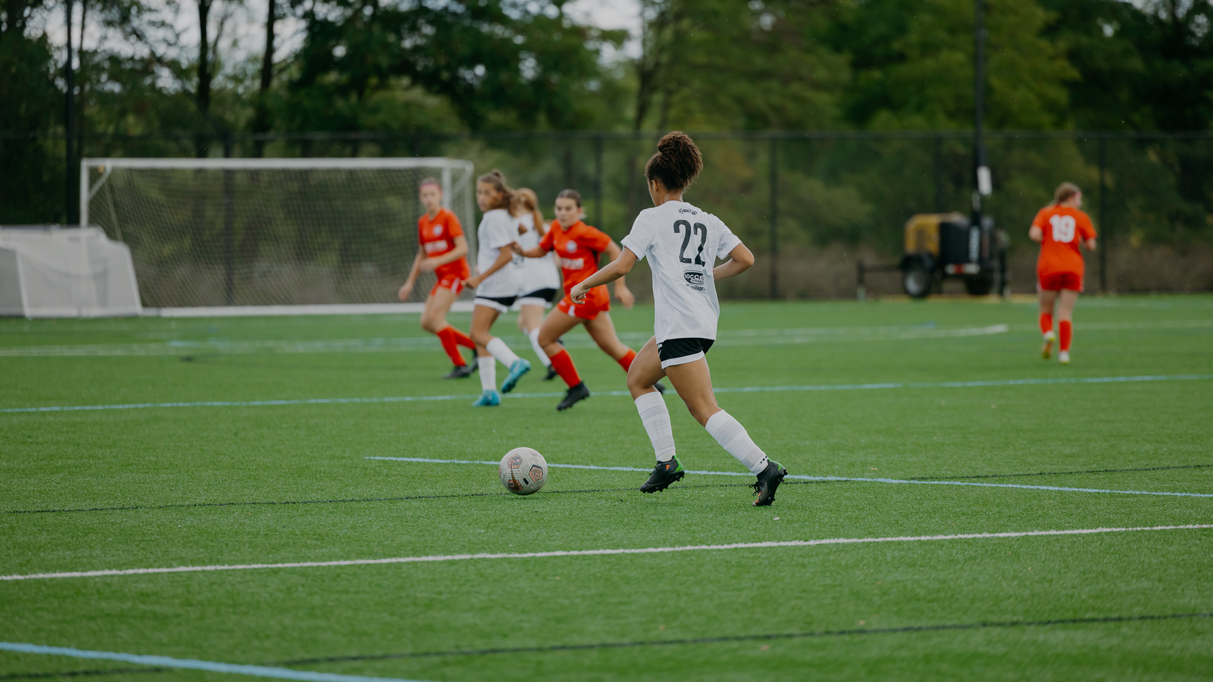 Young women playing soccer on a green field, with one girl in a white jersey kicking the ball and others in red jerseys nearby.
