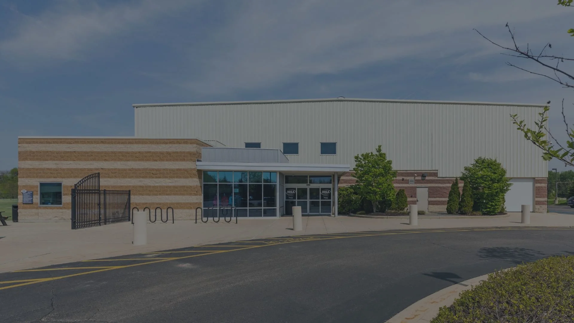 Front view of a modern school building with brick and metal siding, large glass entrance doors, and small trees and shrubs in front.