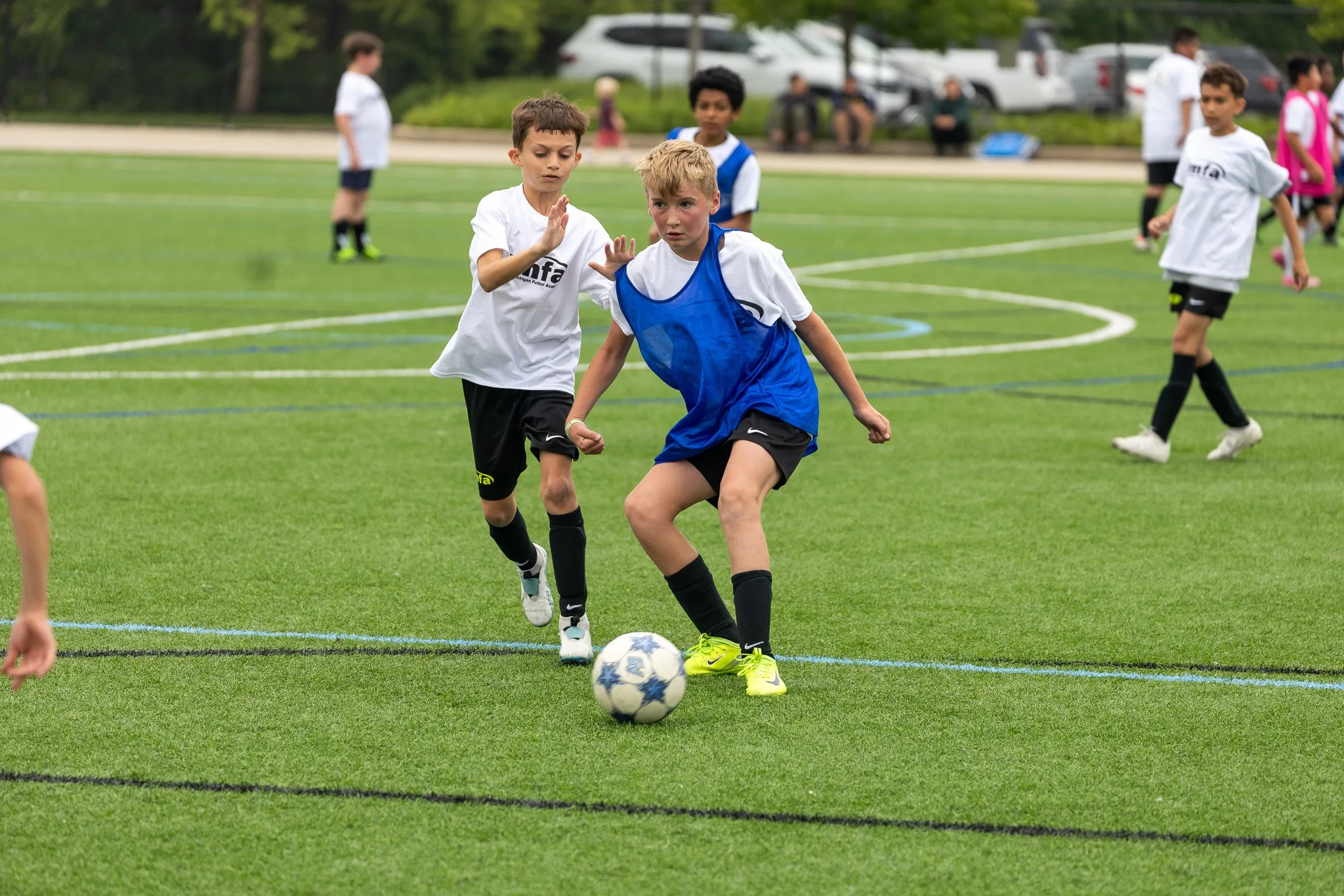 Children playing soccer on a green field during daytime, with some people sitting on the sidelines. Two boys in the foreground are competing for the ball, one in a white shirt and black shorts, the other in a blue vest over a white shirt and dark shorts, both focused on the ball.