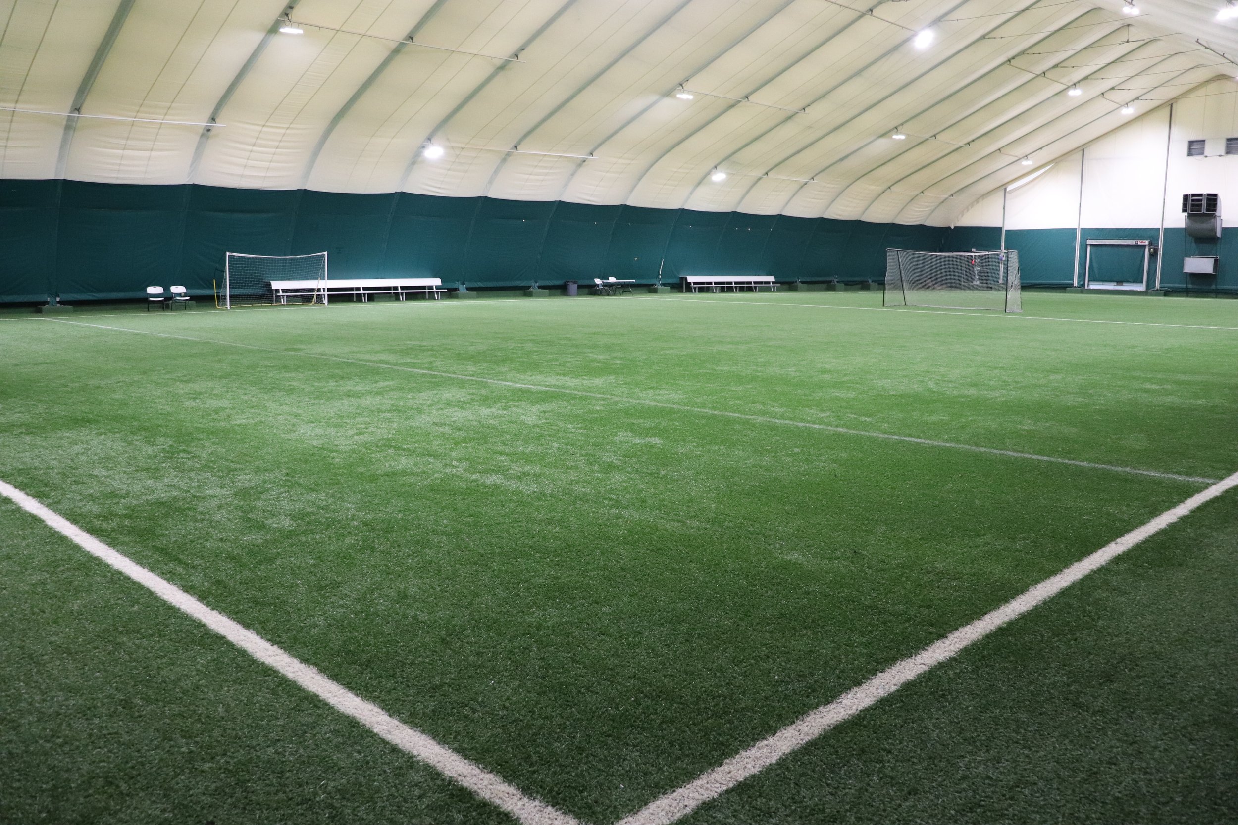 Indoor tennis court with green surface, white lines, and nets, set inside a large dome with lighting and seating along the sides.