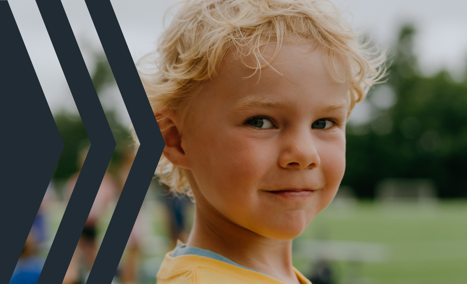 Close-up of a young boy with blonde curly hair and blue eyes, smiling outdoors at a park or sports field.