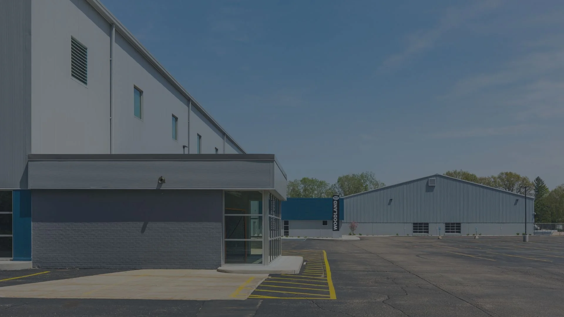 An industrial building with a sign reading 'Woodland' on the side, a parking lot with yellow lines, and a small glass entrance in the foreground. The sky is blue with some clouds, and there are trees in the background.