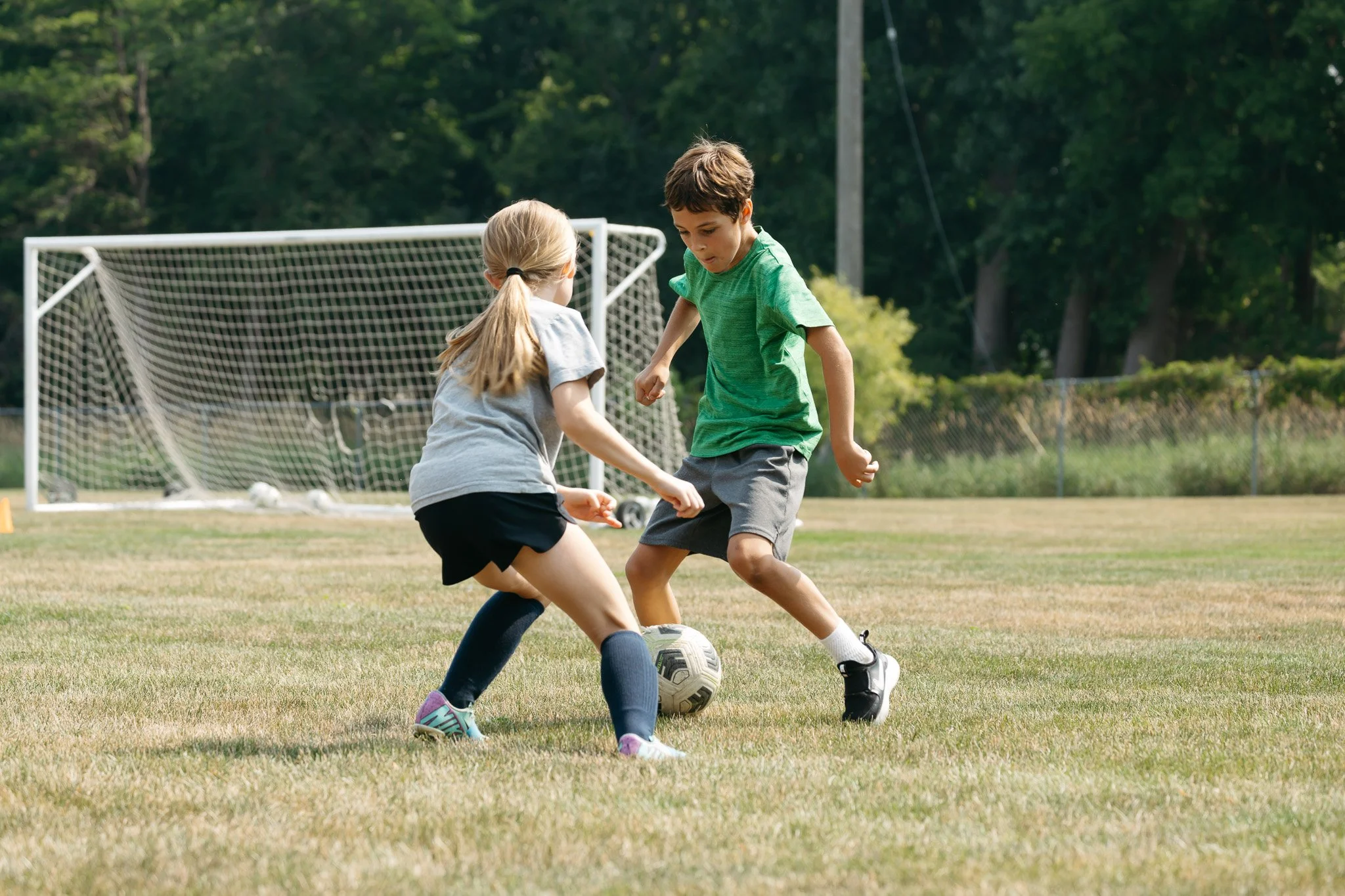 Two children, a girl and a boy, playing soccer on a grassy field with a soccer goal in the background, surrounded by trees.