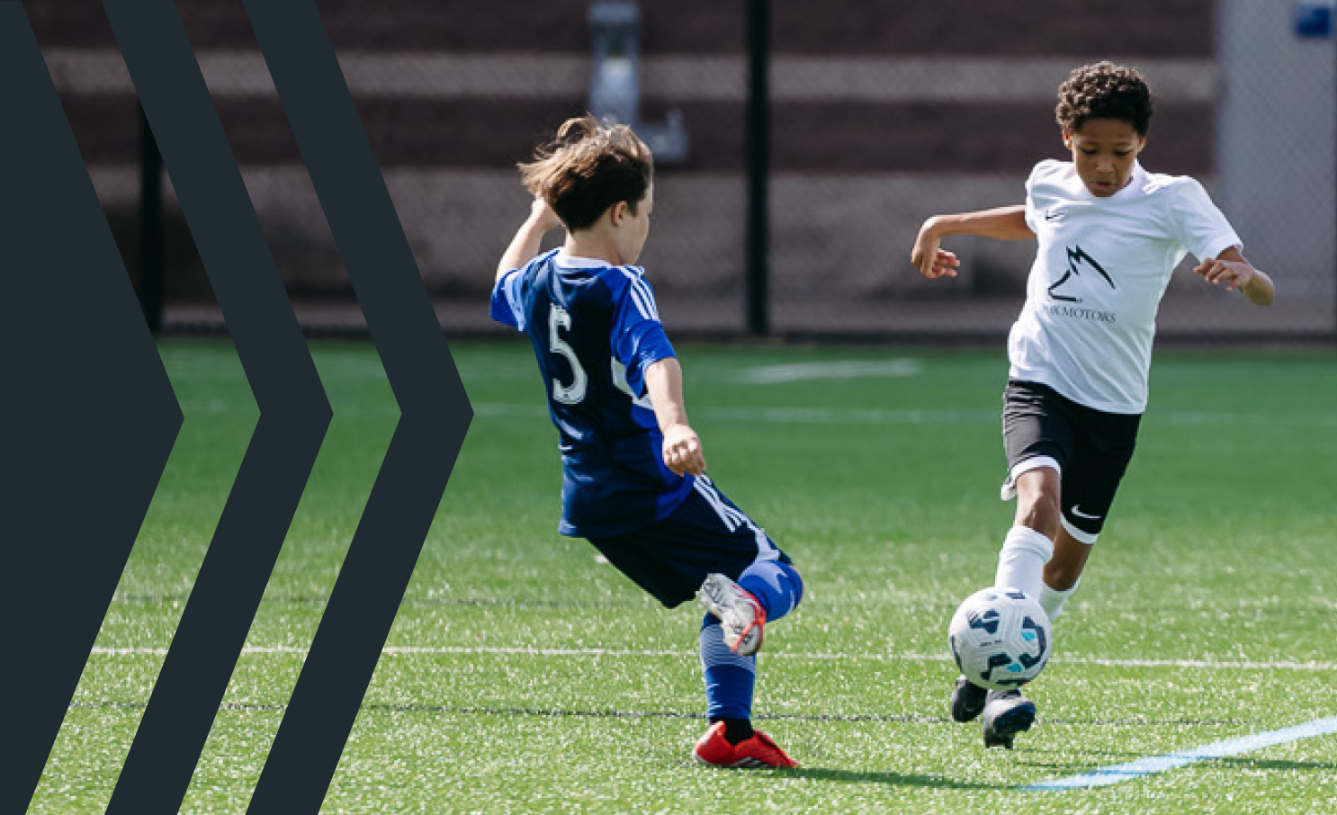 Two young boys playing soccer on a field, one in a white shirt and black shorts kicking the ball, the other in a blue jersey and shorts approaching the ball.