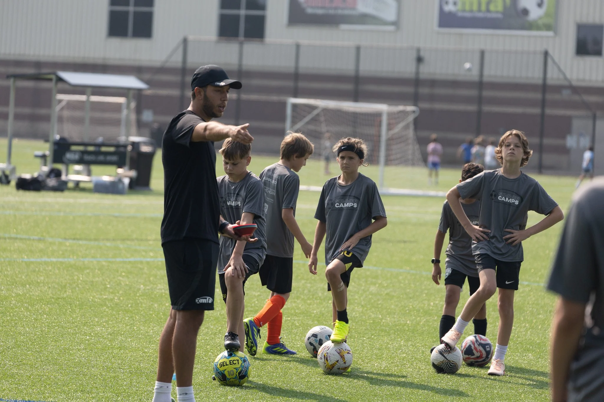 A coach instructs young soccer players during a practice session on a grassy field. The players are wearing gray shirts, some with black headbands, and are standing near soccer balls. In the background, there are goalposts, a fence, and a building.