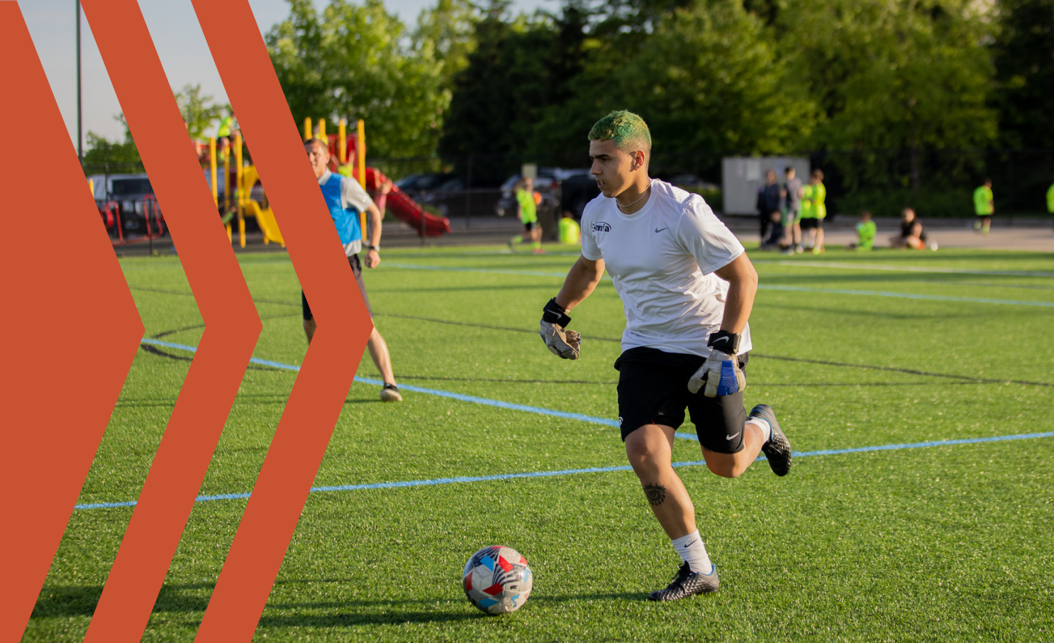A person with green hair wearing a white shirt and black shorts playing soccer on a grassy field, with children and adults in the background.