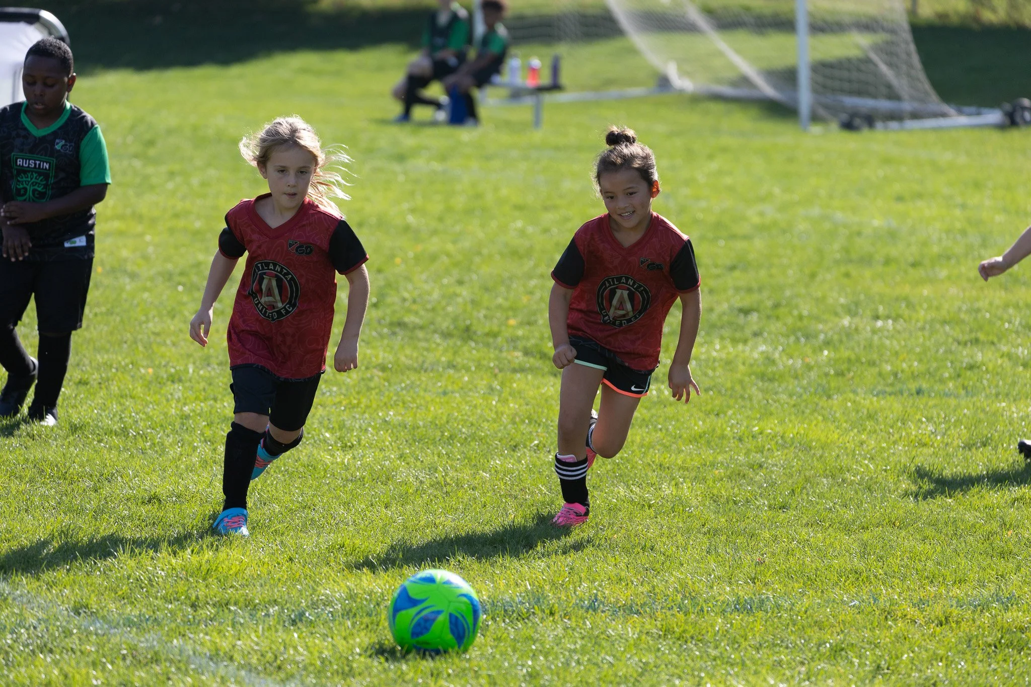 Children playing soccer on a grassy field, with two girls running towards a green and blue ball in the foreground, and two boys in the background wearing green and black uniforms.