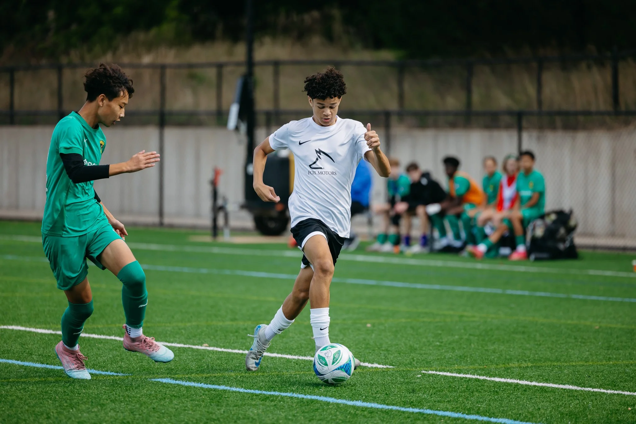 Two young soccer players, one in a white jersey and the other in a green jersey, competing for the ball on a soccer field. A group of players sits on a bench in the background.