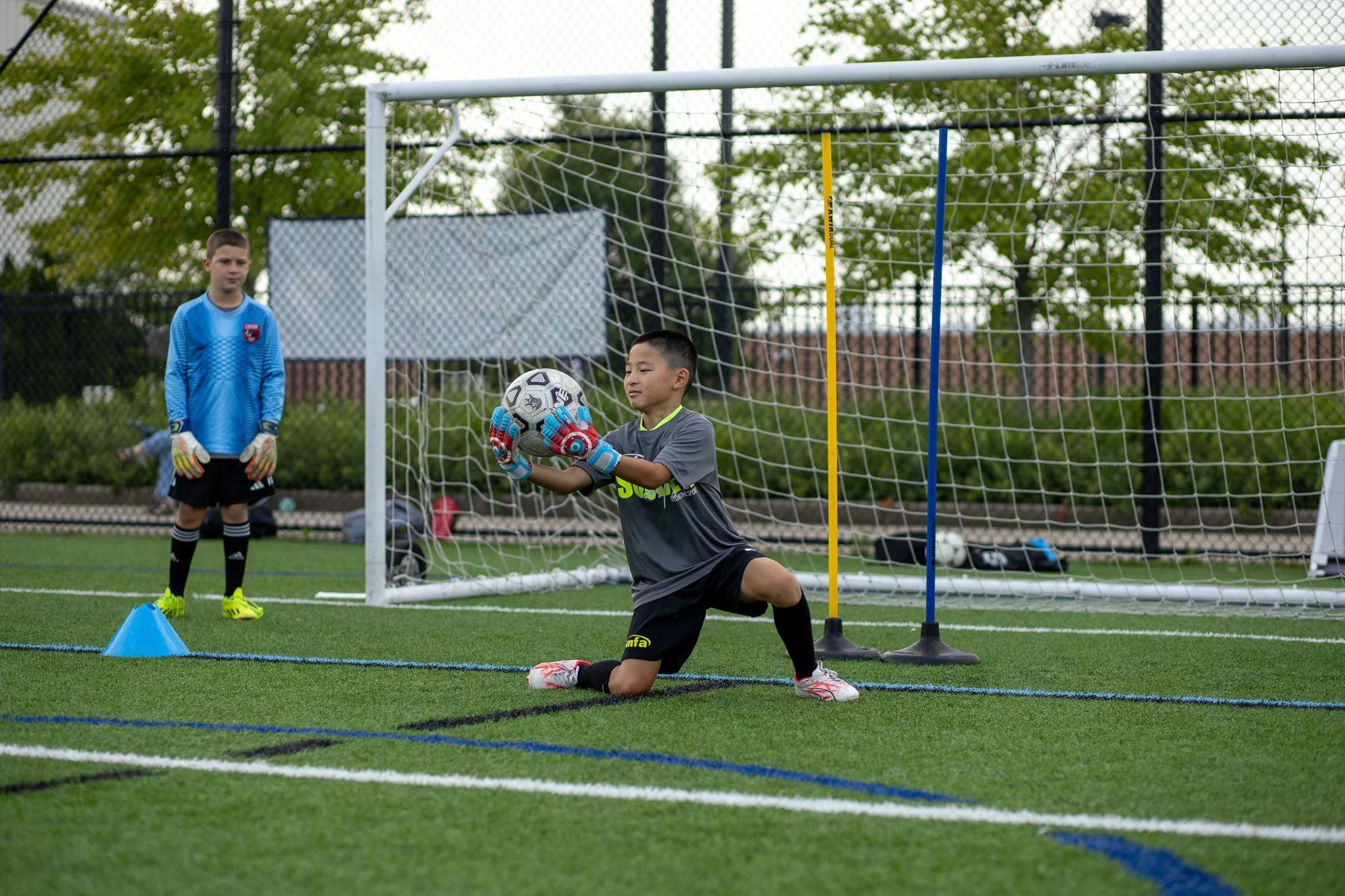 Young children playing soccer on a field, with one kid wearing gloves and holding a black and white soccer ball, kneeling near the goal, and another kid standing nearby.
