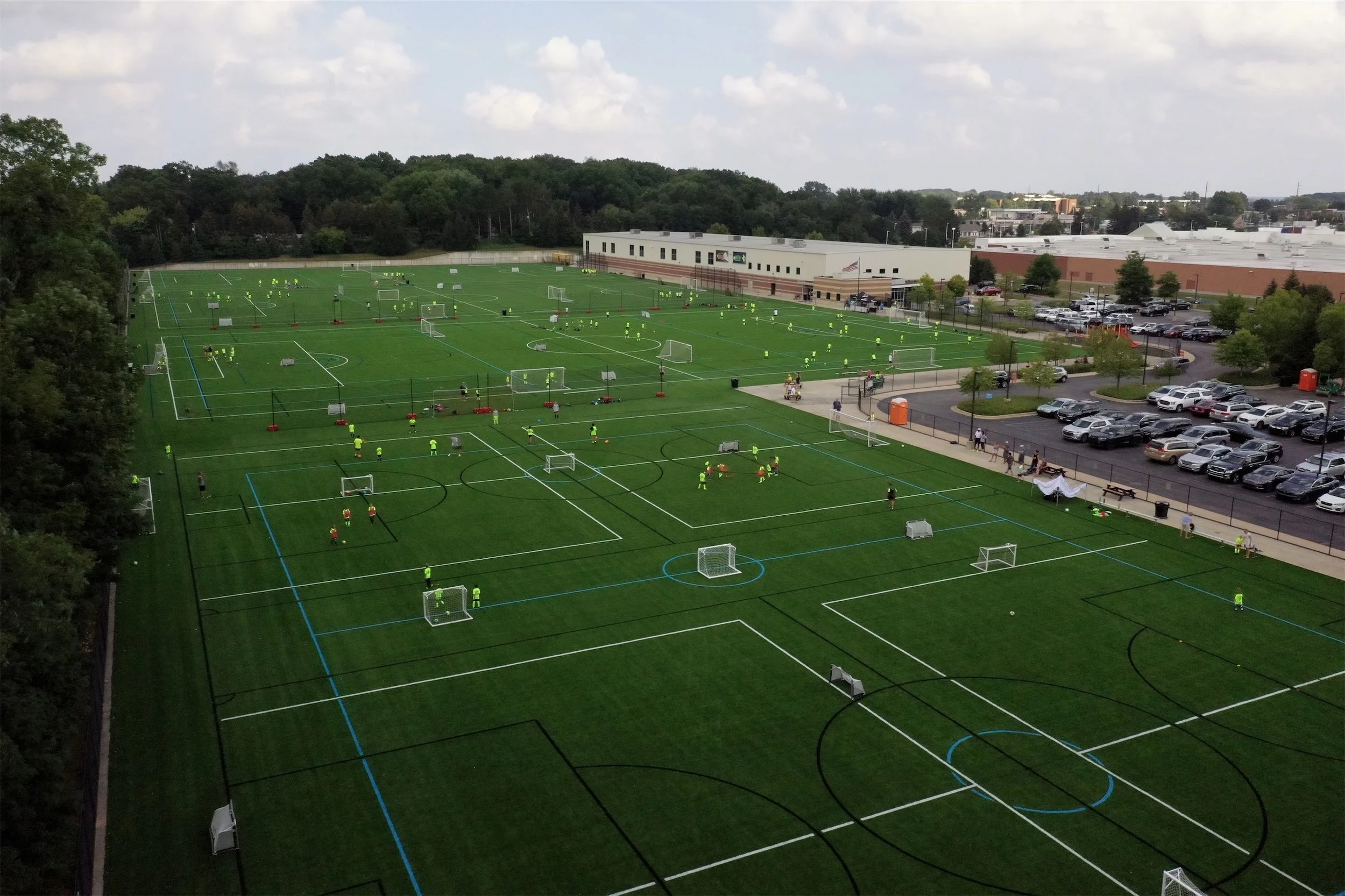 Multiple artificial turf soccer fields with players practicing and playing, adjacent to a parking lot and a school building, under a partly cloudy sky.