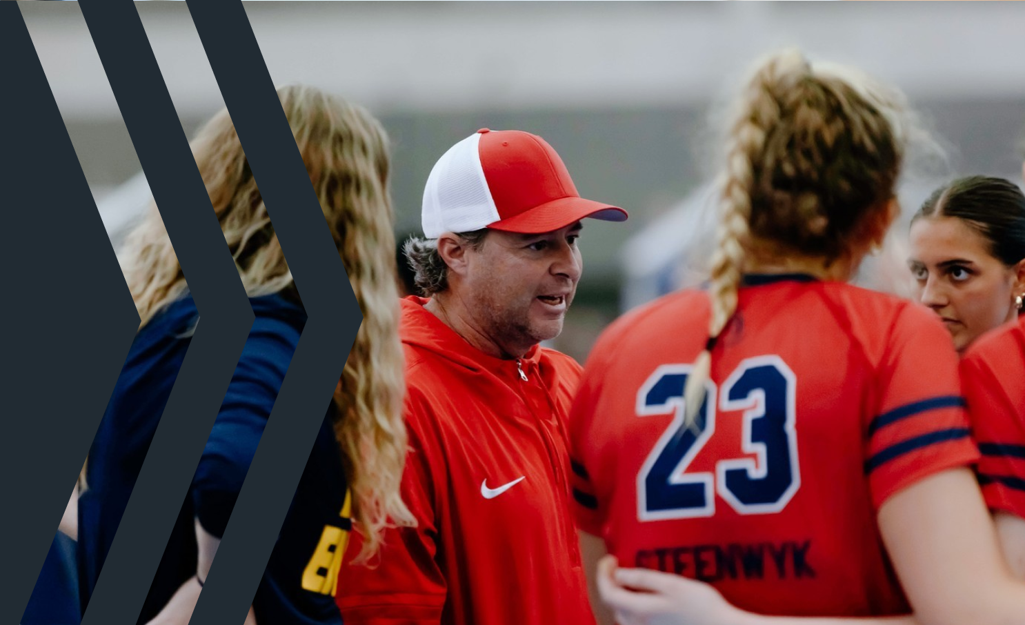A coach speaking to female athletes on a sports field, wearing red and navy uniforms with the number 23 visible.