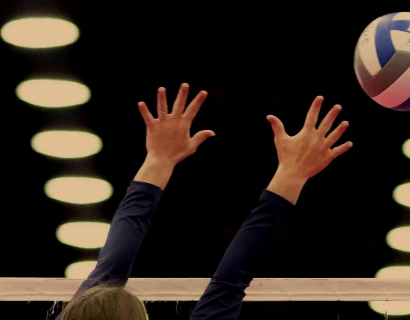 A person with long hair and a dark long-sleeve shirt reaching up with two hands to hit a colorful volleyball in a dimly lit indoor setting.