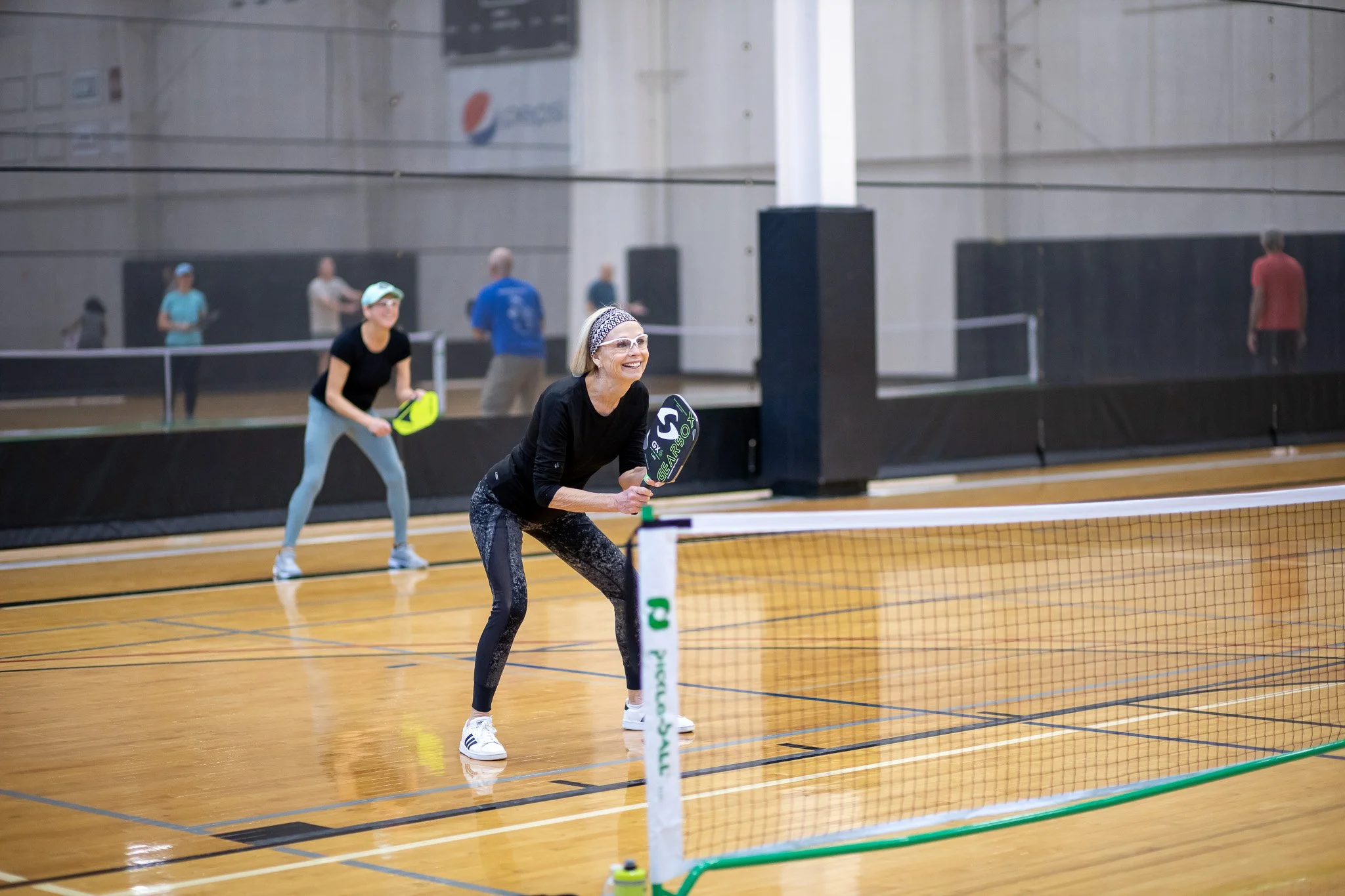 Women playing pickleball indoors, smiling and holding paddles near a net on a wooden court.