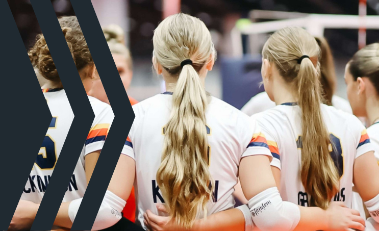 Group of female volleyball players huddled together during a match, seen from behind, wearing white jerseys with colorful accents and long hair tied back.