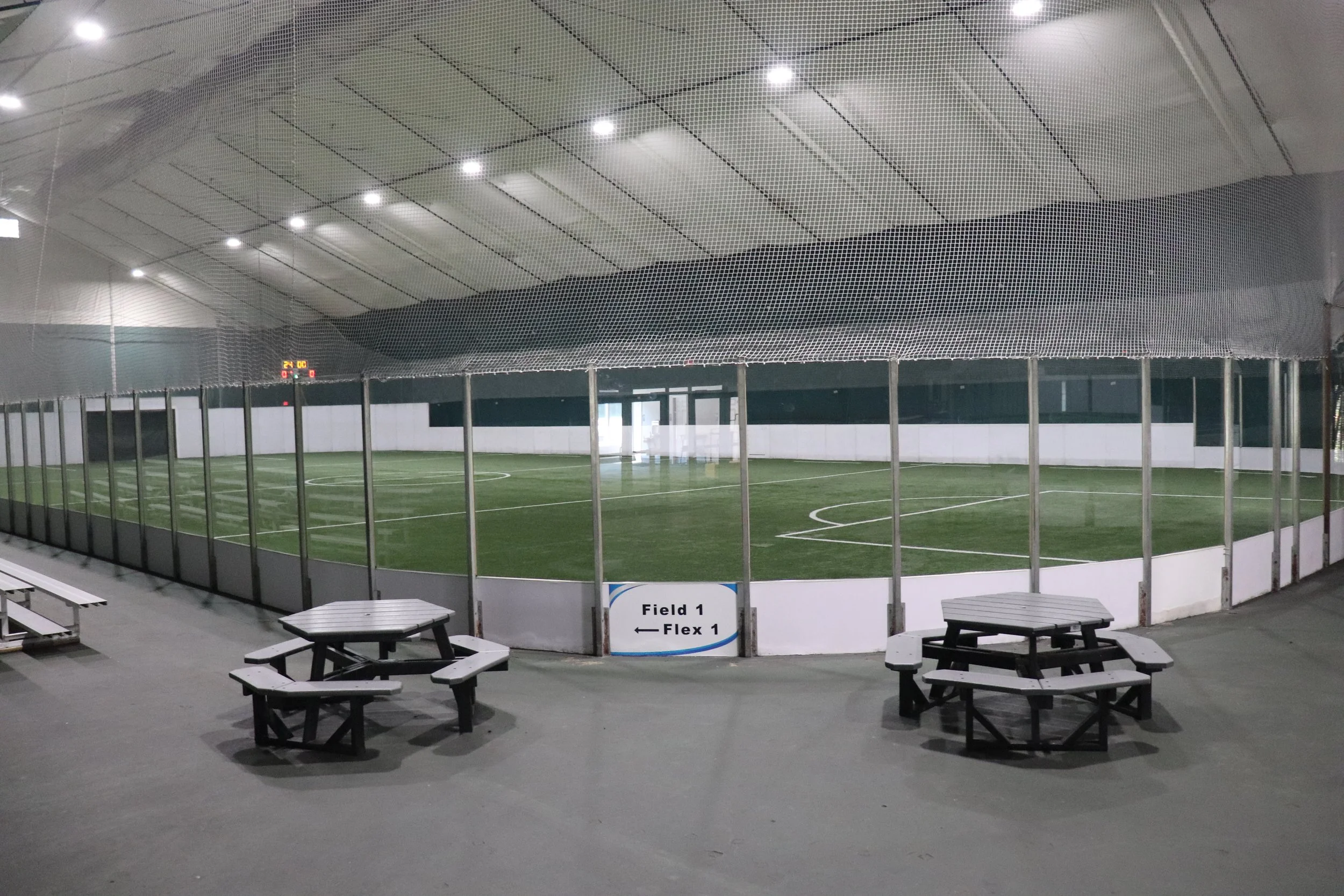 Indoor sports arena with artificial turf field, surrounded by glass panels and a netted ceiling. There are benches and signs indicating Field 1 and Flex 1.