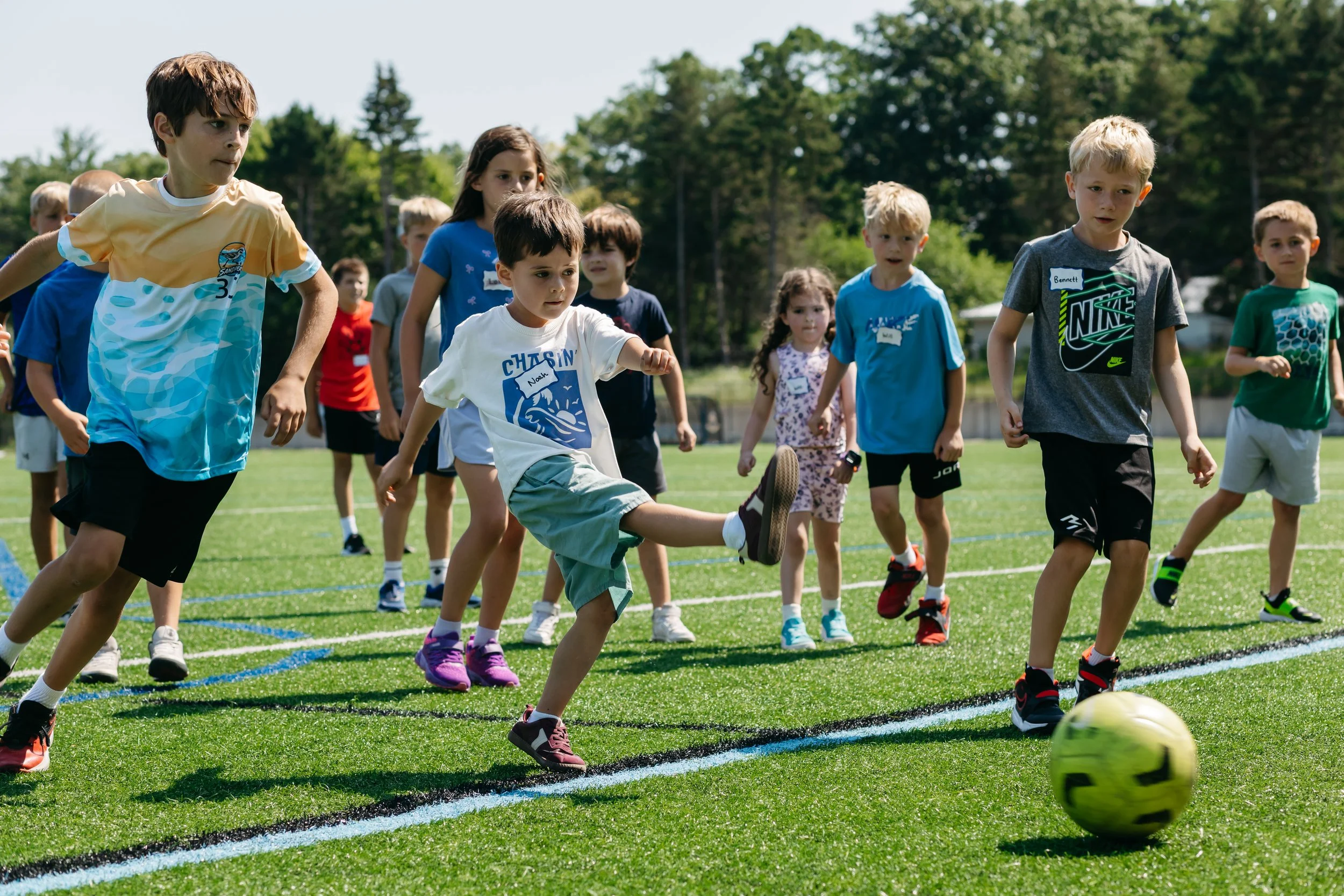 Children play a soccer game during All Day with MSA at Michigan Sports Academies, with a young camper kicking a soccer ball while other kids run across the turf field. The youth sports camp in West Michigan focuses on development and teamwork.