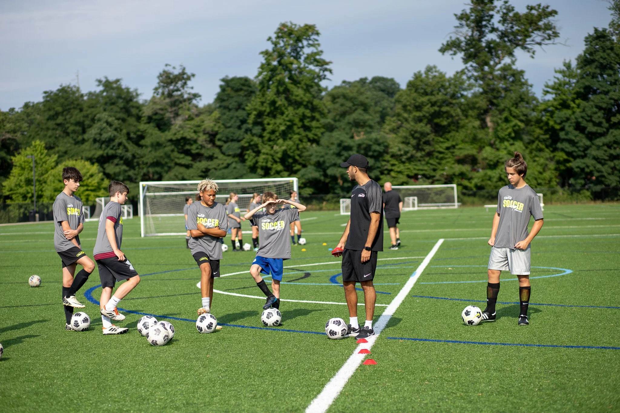 A youth soccer practice on a field with young players, coaches, and soccer balls, with trees and soccer goals in the background.