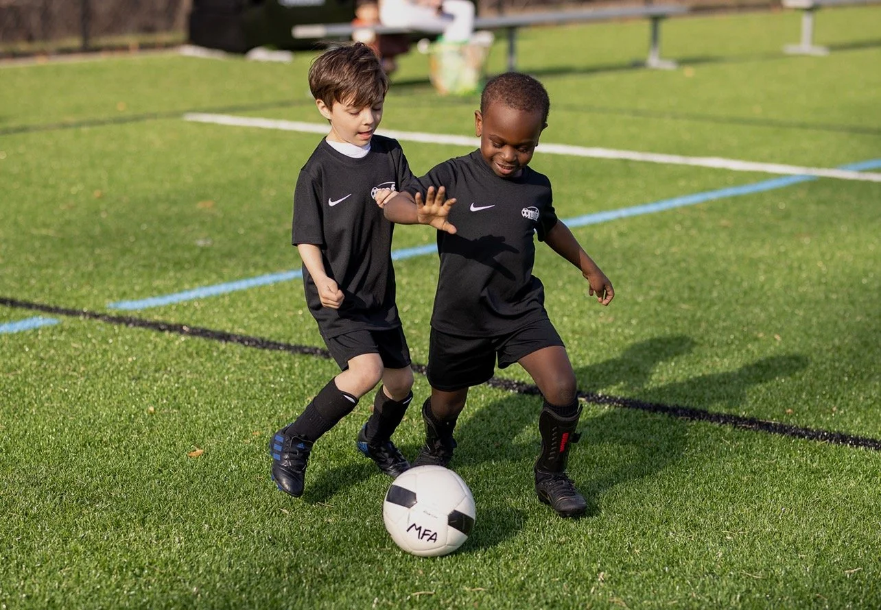 Two young boys in black soccer uniforms playing soccer on a field, one trying to kick the ball while the other attempts to block.