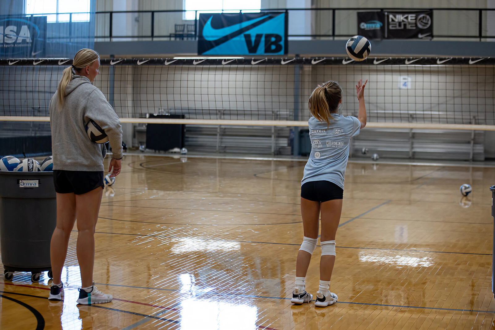 Two young women playing volleyball indoors, one preparing to serve the ball and the other watching, in a gym with a volleyball net and banners hanging on the wall.