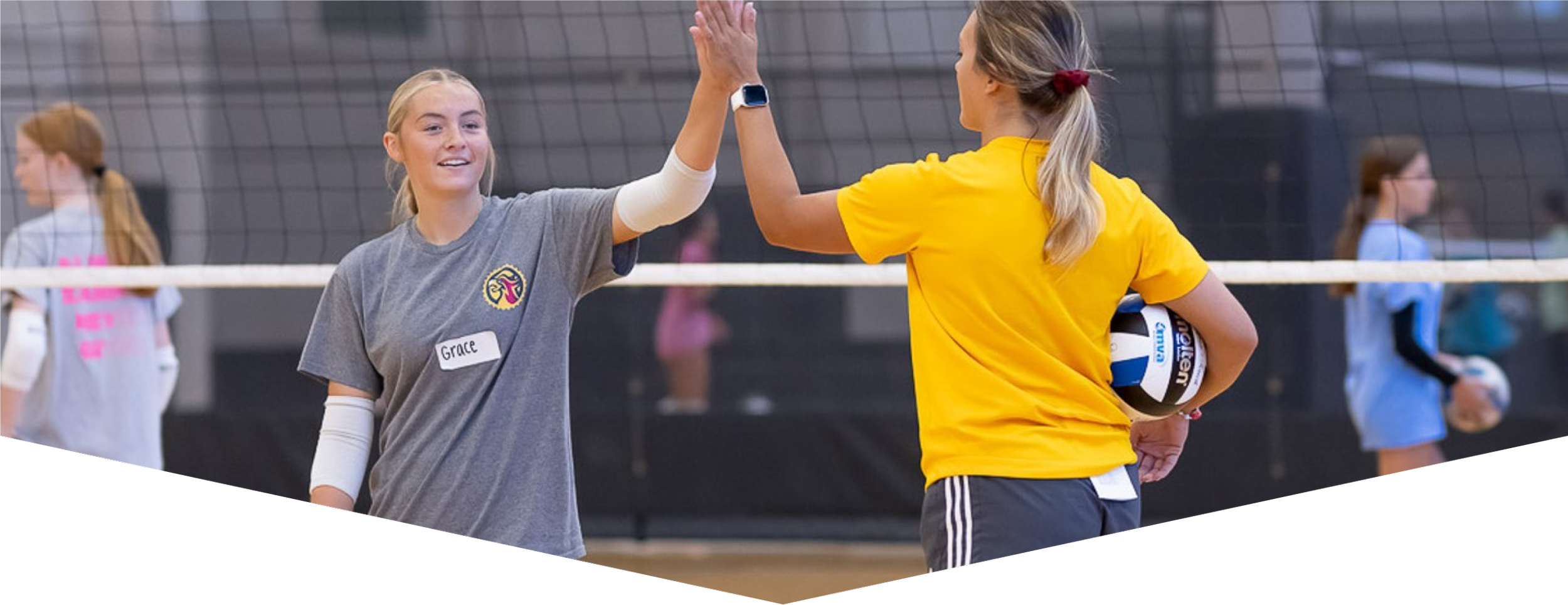 Two young women high-fiving during a volleyball game in an indoor gym. One woman is holding a volleyball, and the other is wearing a gray shirt with a name tag that reads "Grace." Others are present in the background.