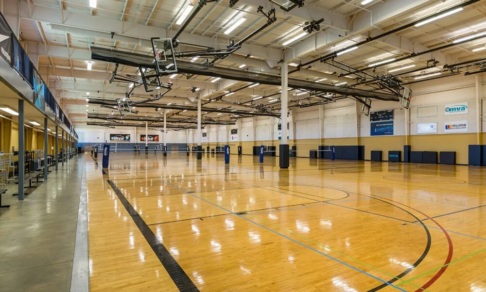Empty indoor volleyball court with polished wooden floor, overhead basketball hoops, and padded walls in a gymnasium.