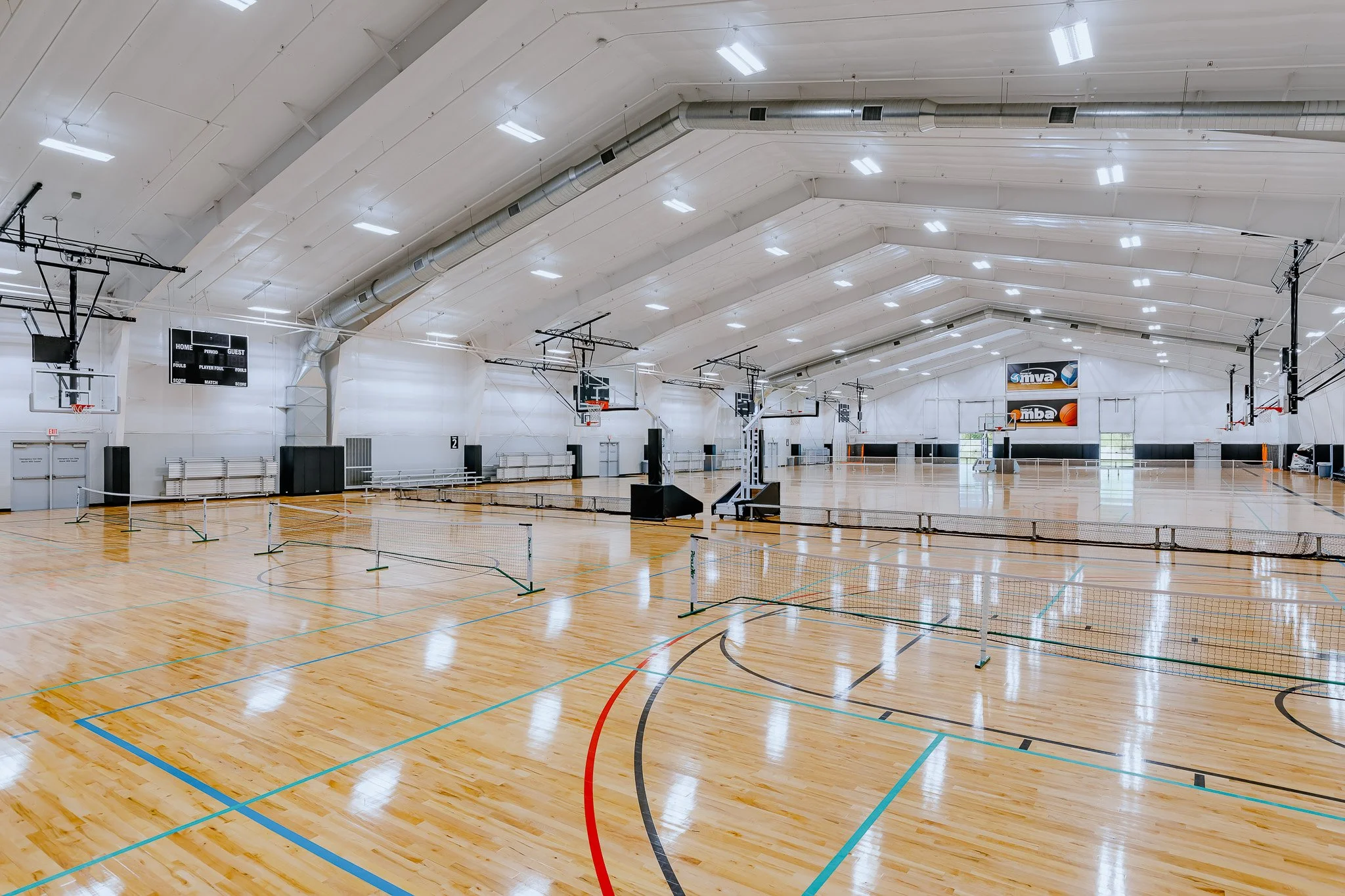 An indoor basketball gymnasium with a wooden floor, multiple basketball hoops, scoreboards, and a badminton net set up. The ceiling is high with bright lighting and large ventilation ducts.