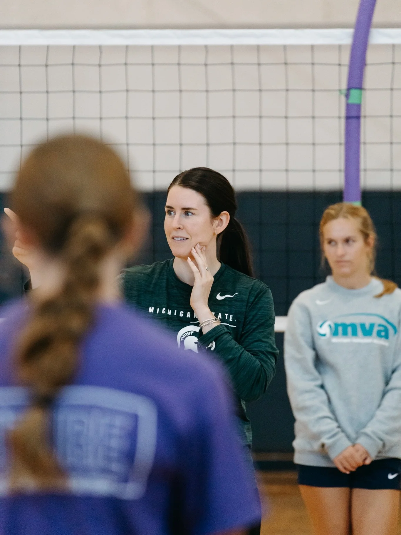 A volleyball coach or instructor speaking to a group of young female volleyball players in a gymnasium. The coach is wearing a Michigan State university shirt, and the players are in sportswear, with one in a gray shirt and another in a purple jersey. Volleyball net in the background.