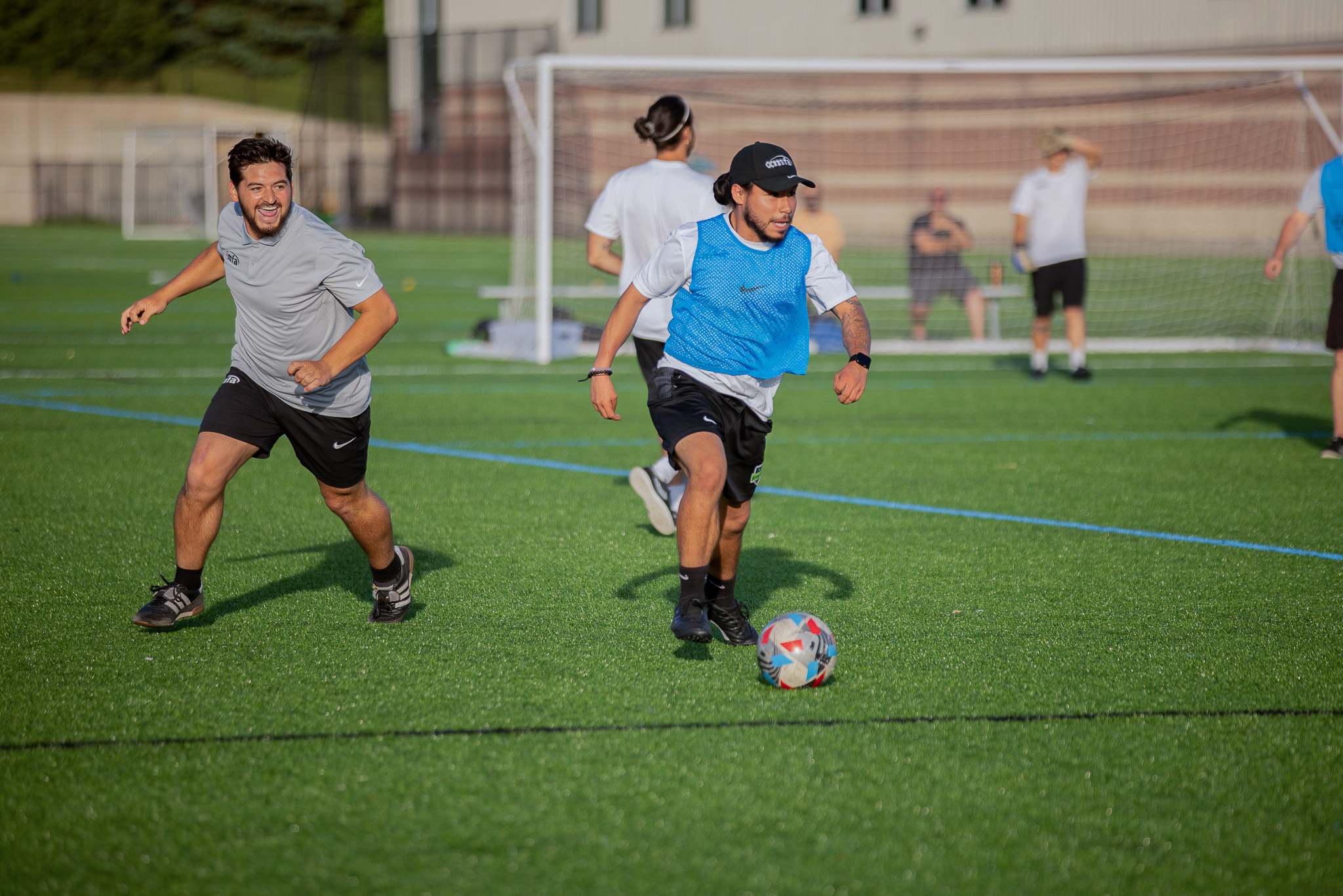 Two men playing soccer on a field during daytime, with others in the background. One man is running towards the ball while the other is trailing behind. The overall scene shows a friendly soccer game with people in casual athletic attire.