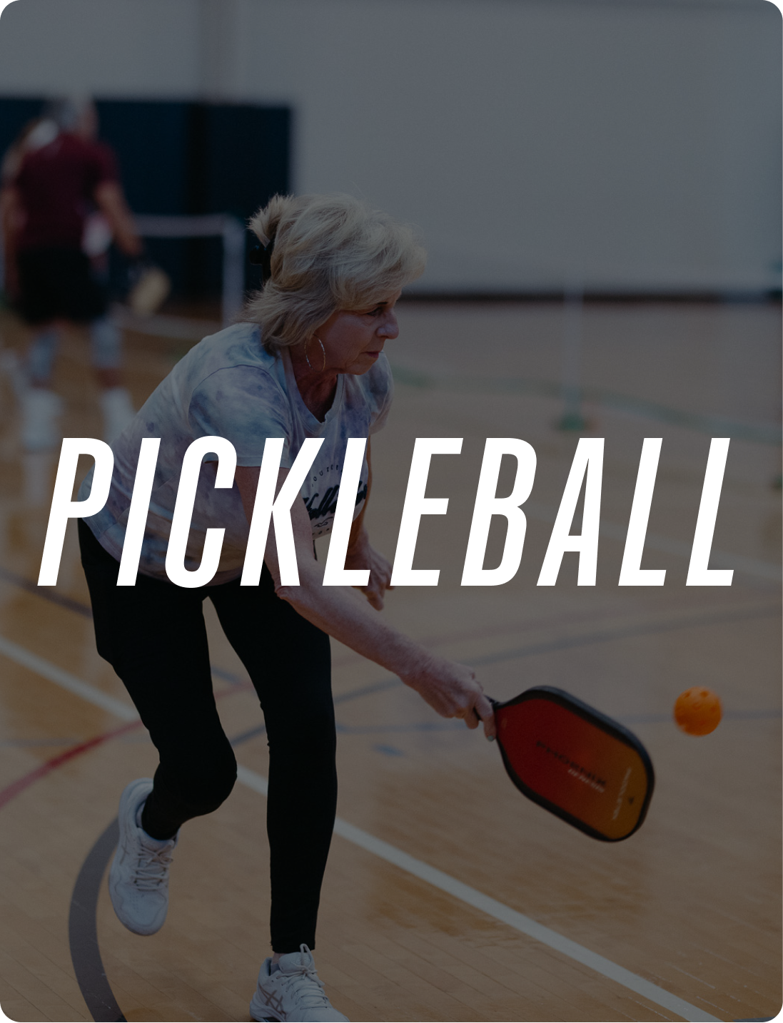 An older woman playing pickleball on an indoor court, hitting an orange ball with a paddle.
